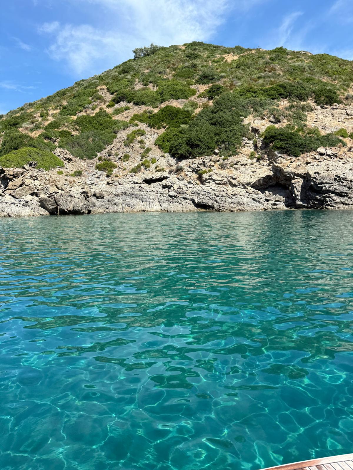 Sparkling turquoise water by a rugged rocky coast and green hillside.