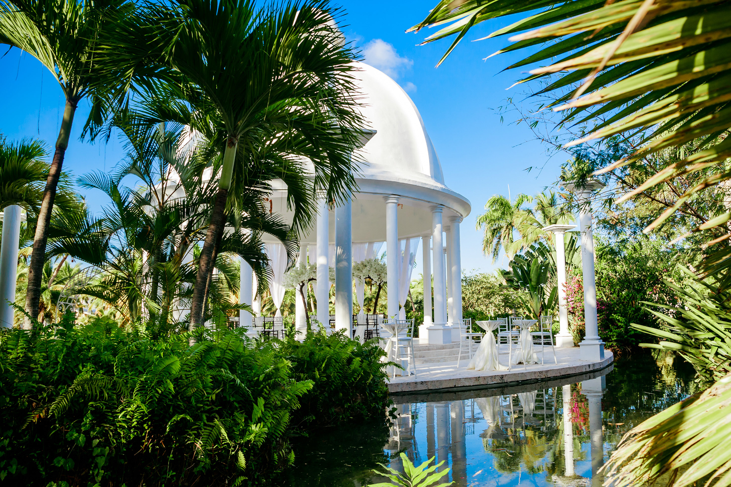 a white gazebo with white columns and a white dome with chairs and a pool of water