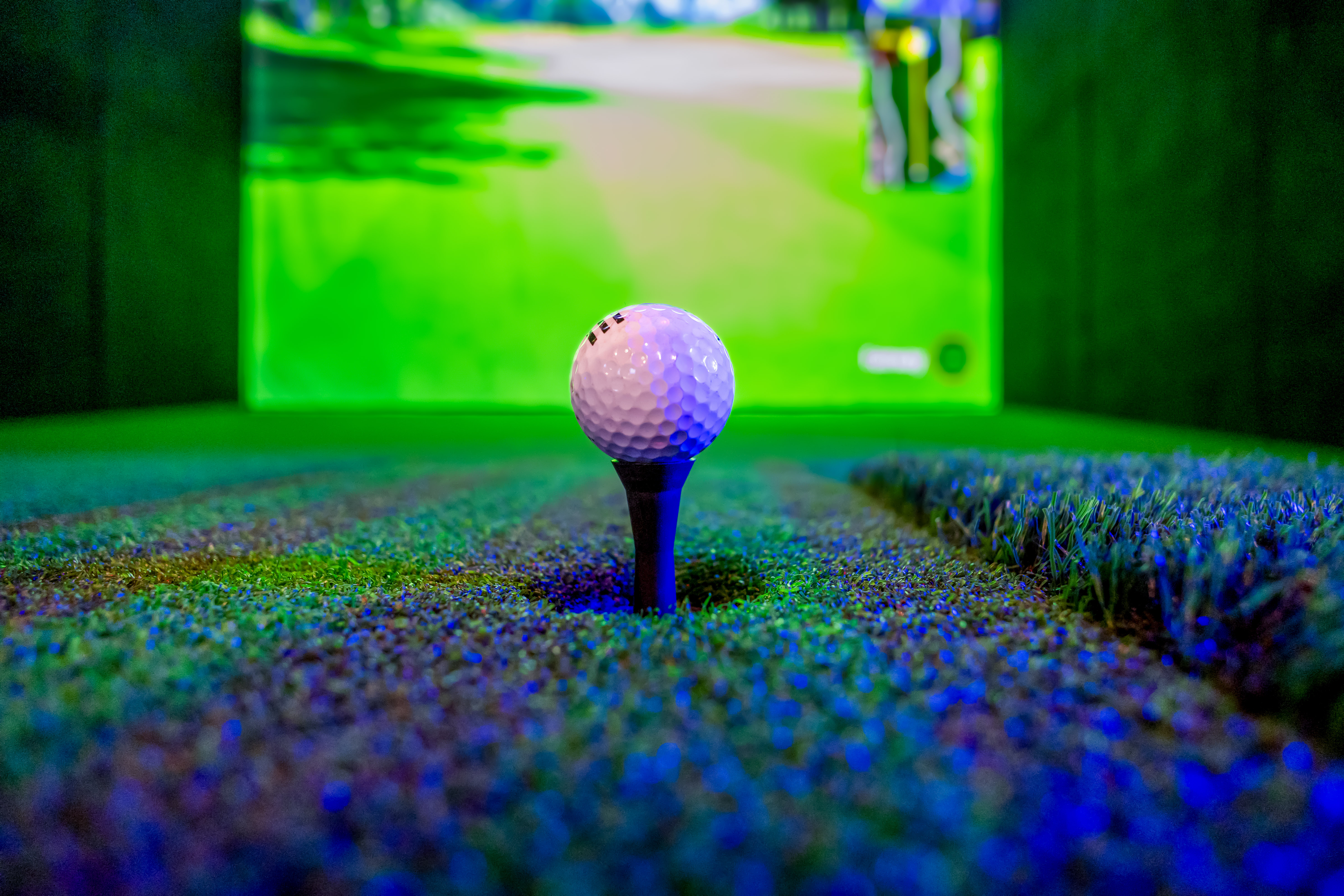 White golf ball on tee, indoor golf simulator with green course screen.