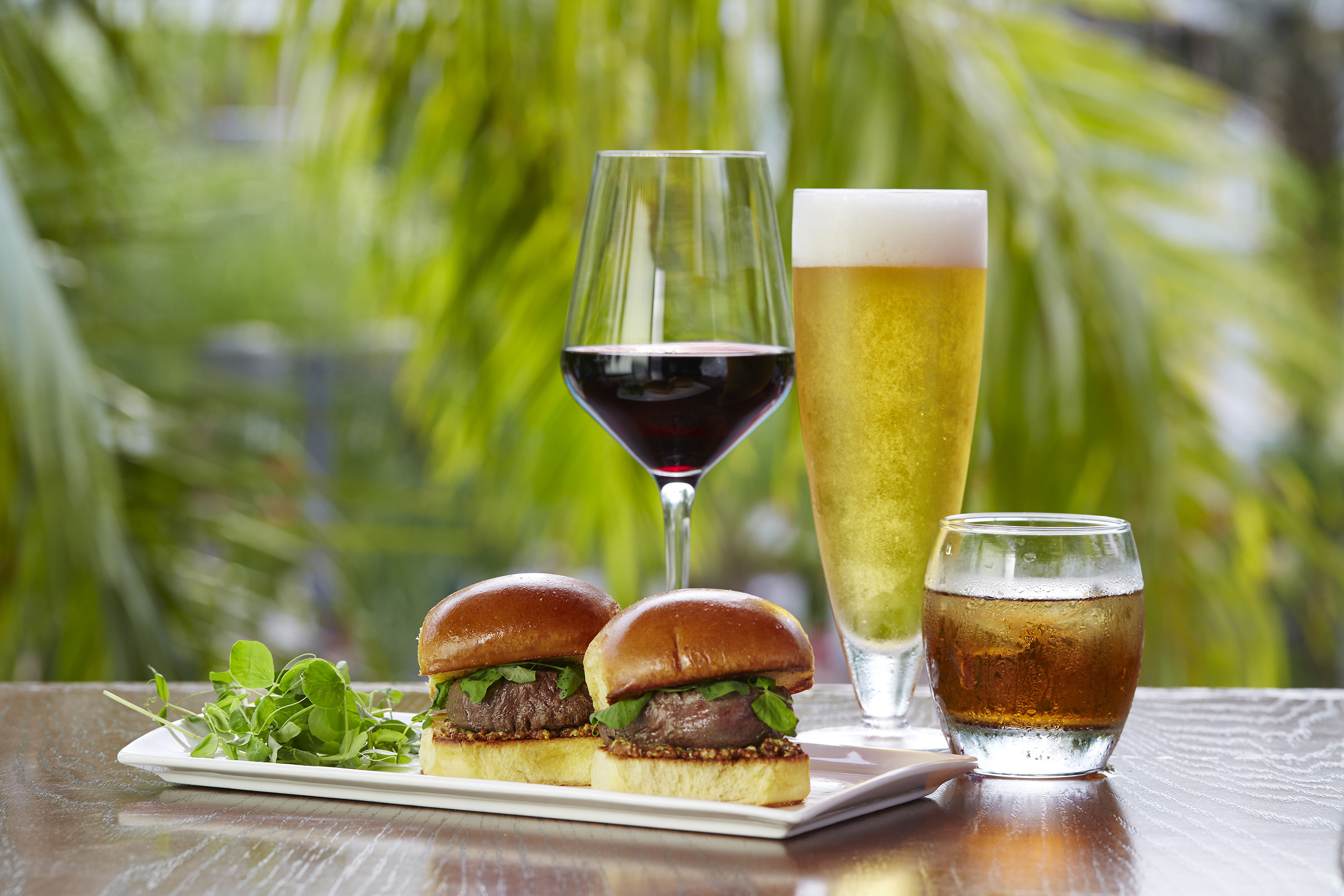 Two gourmet sliders, red wine, beer, and iced drink on an outdoor table.