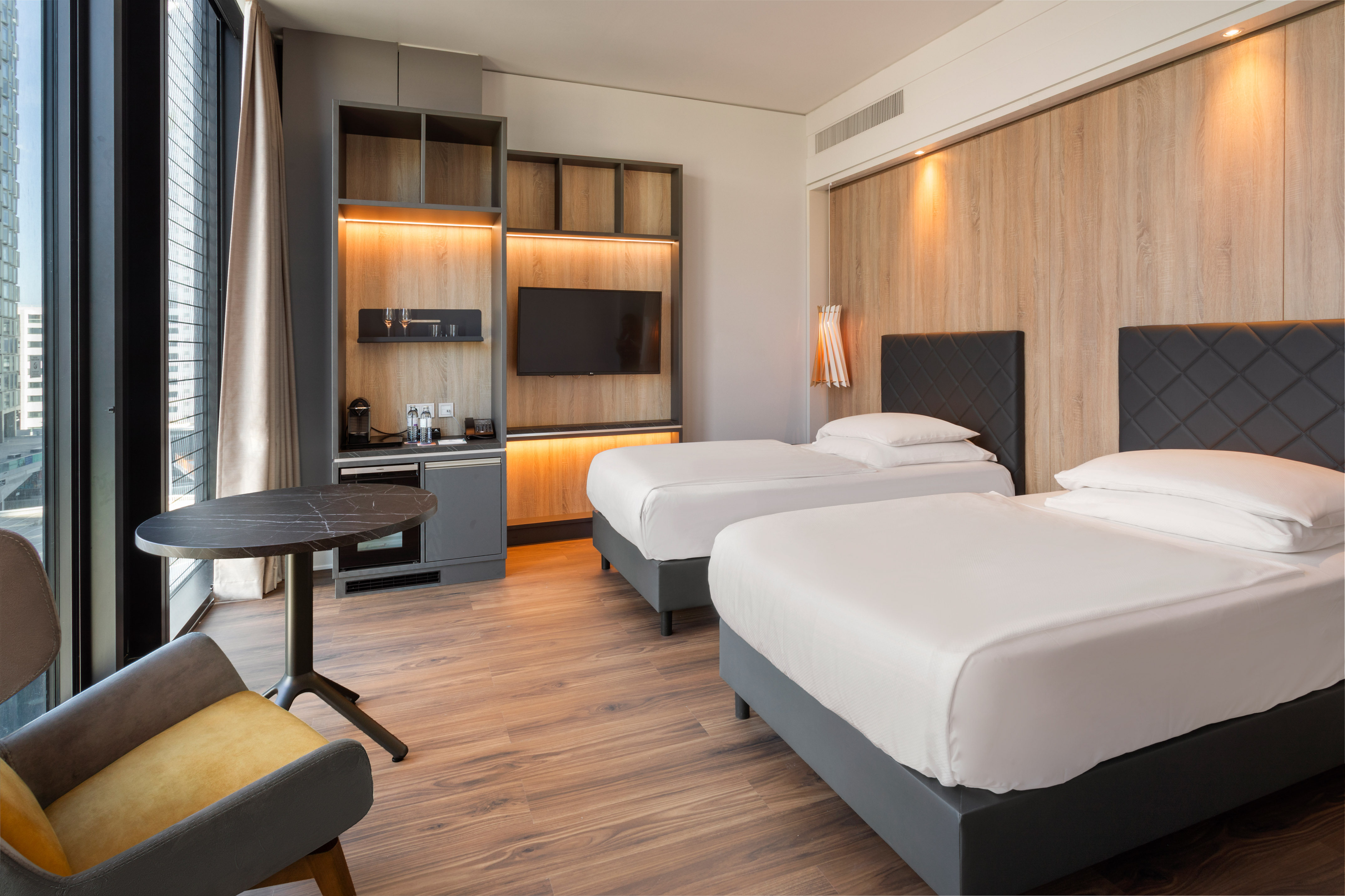 Modern hotel room with two twin beds, city view, wood finishes, and sleek decor.