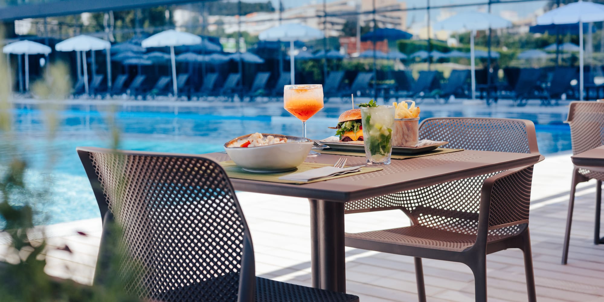 Poolside table set with burger, fries, pasta, and two cocktails.