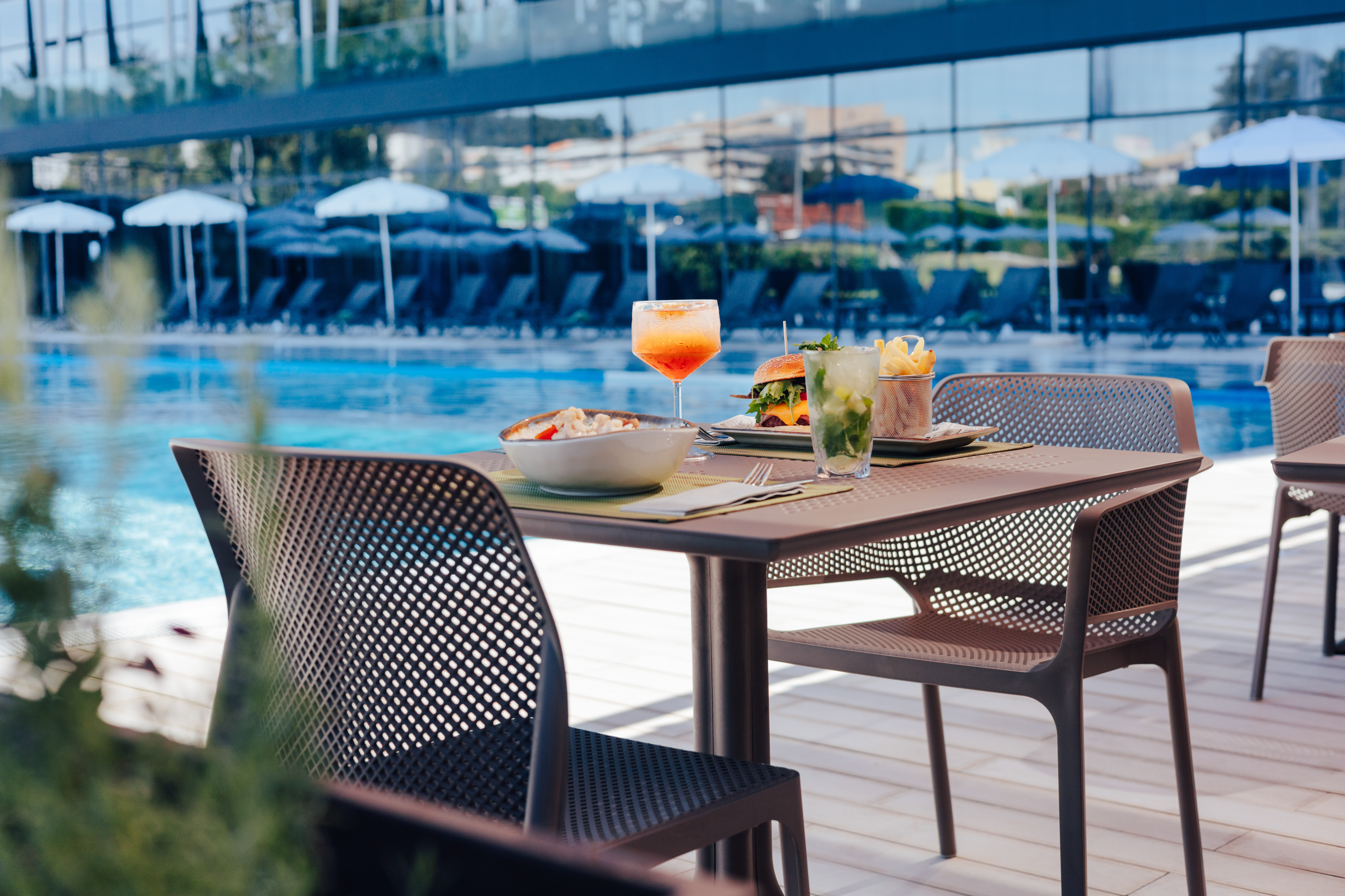 Poolside table set with burger, fries, pasta, and two cocktails.