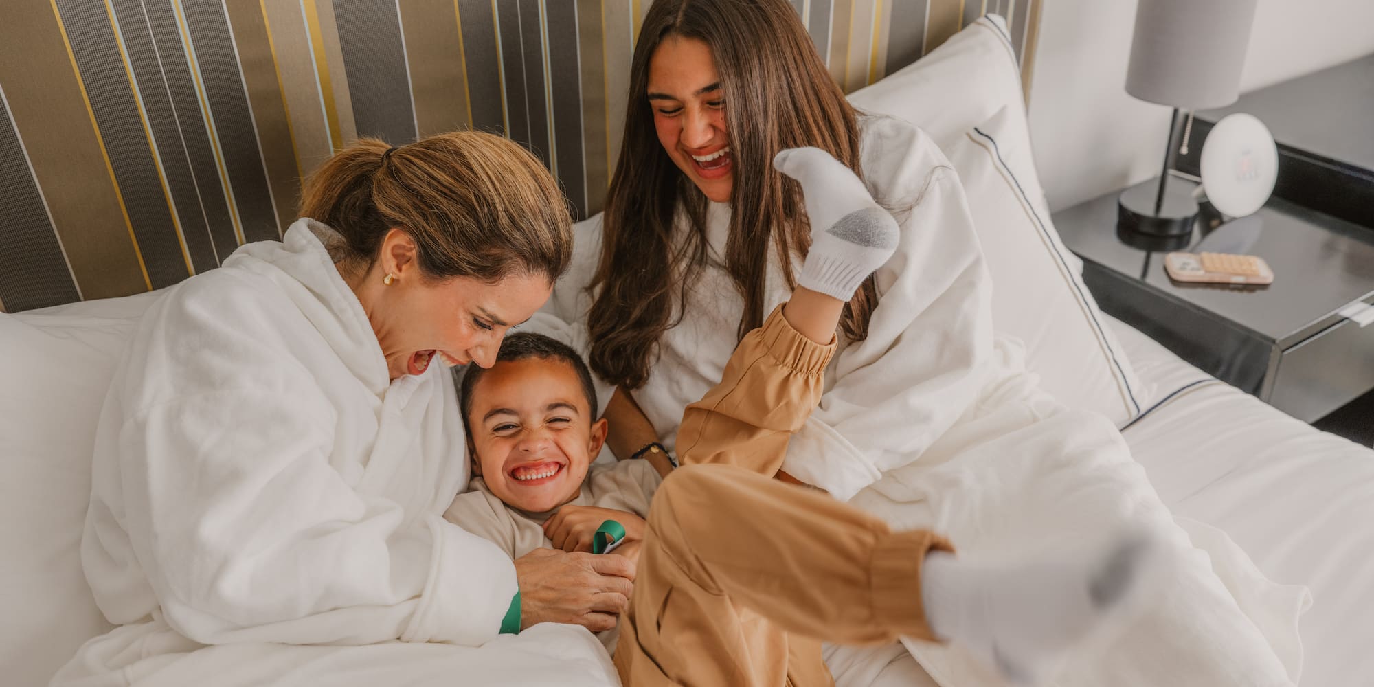 Two women and child laughing, playing in hotel bed, family vacation fun.