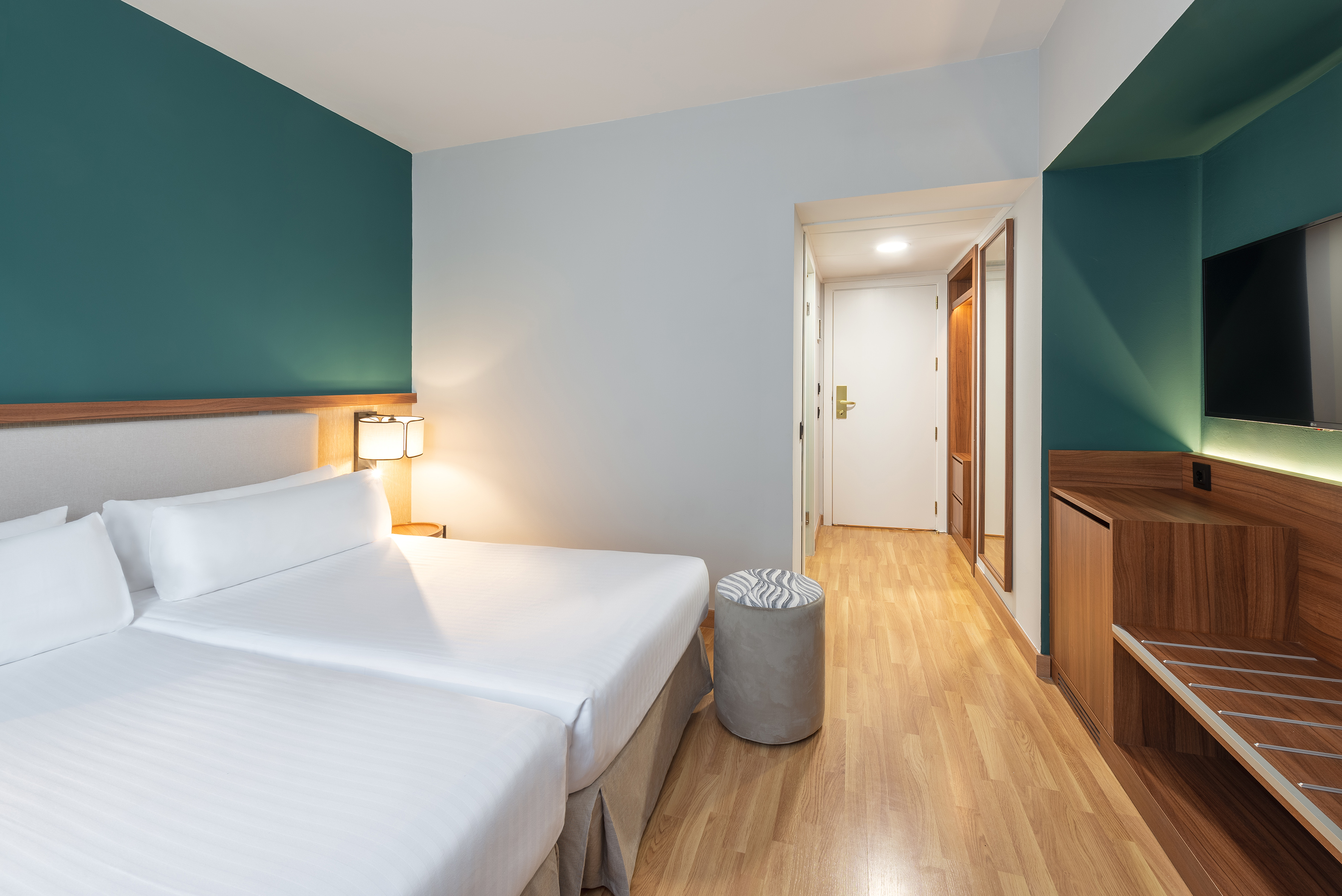 Modern hotel bedroom with twin beds, teal accent wall, and wooden furniture.