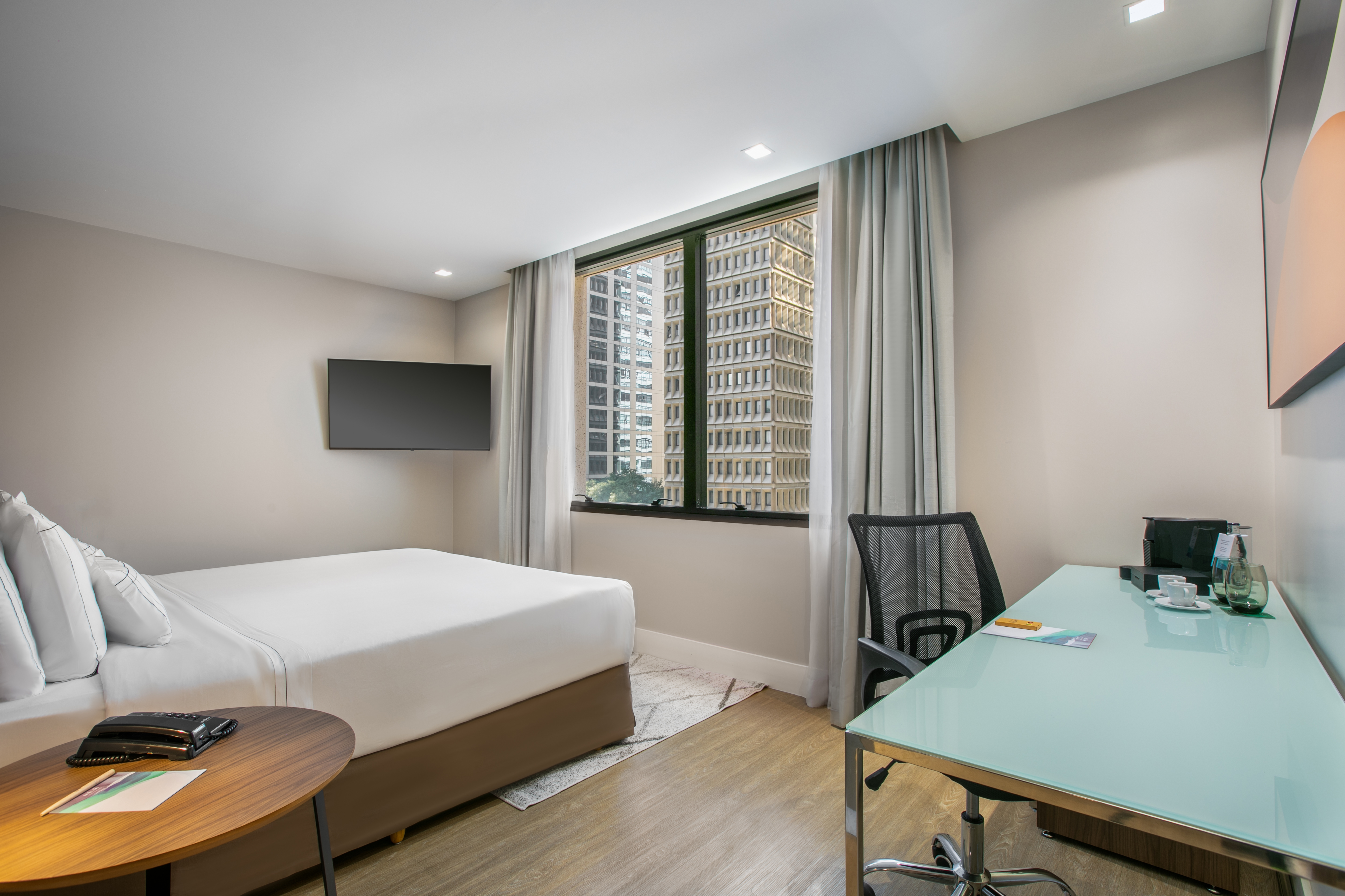 Modern hotel room with bed, desk, wall TV, and urban city view from window.