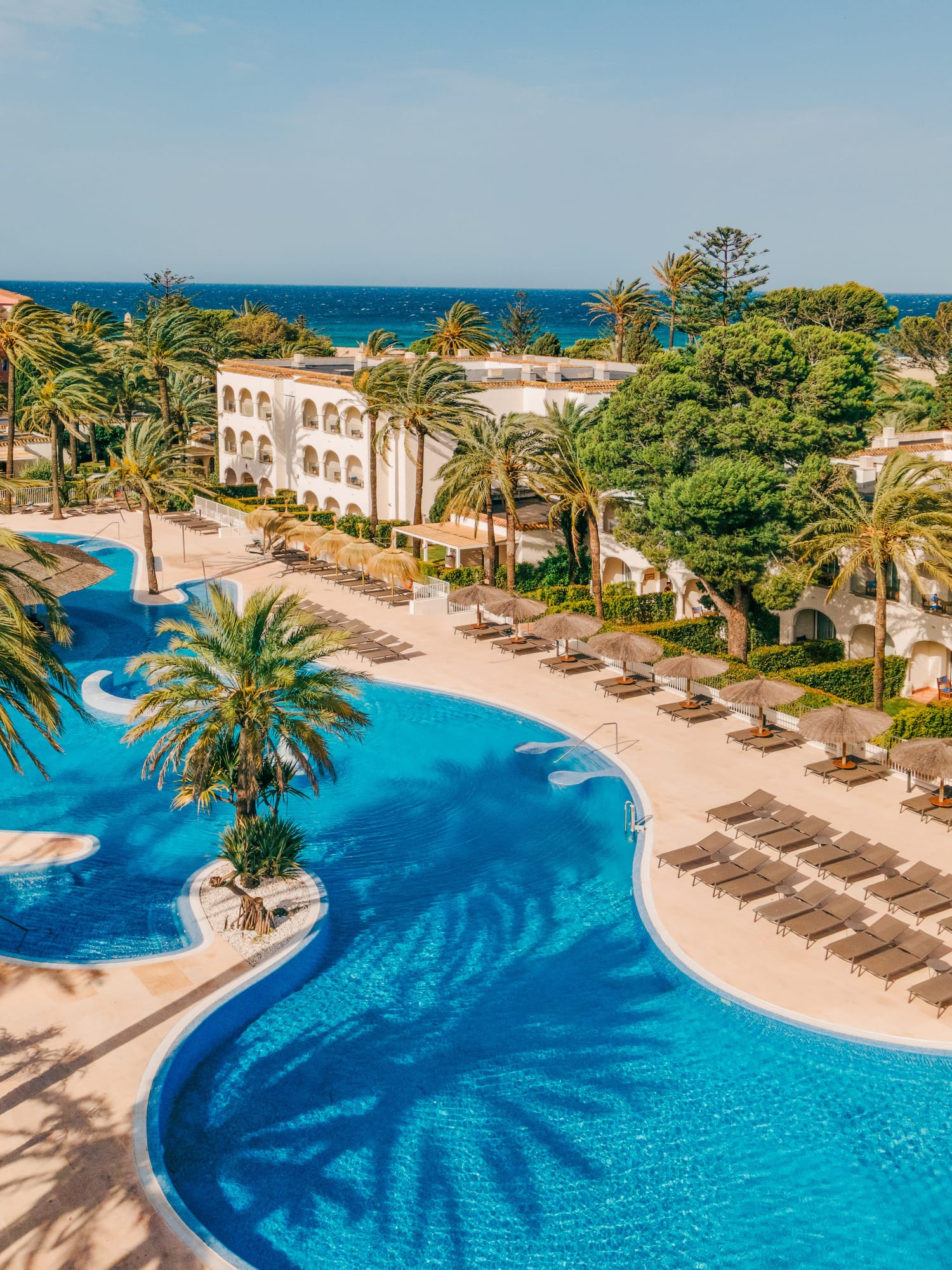 Resort hotel with winding blue pool, palm trees, sun loungers, ocean view.