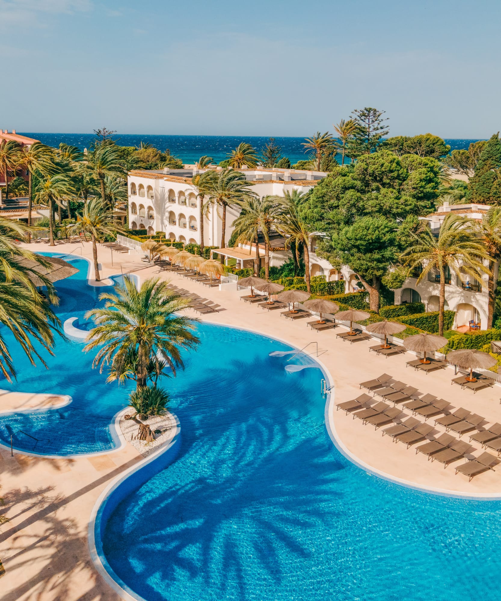 Resort hotel with winding blue pool, palm trees, sun loungers, ocean view.
