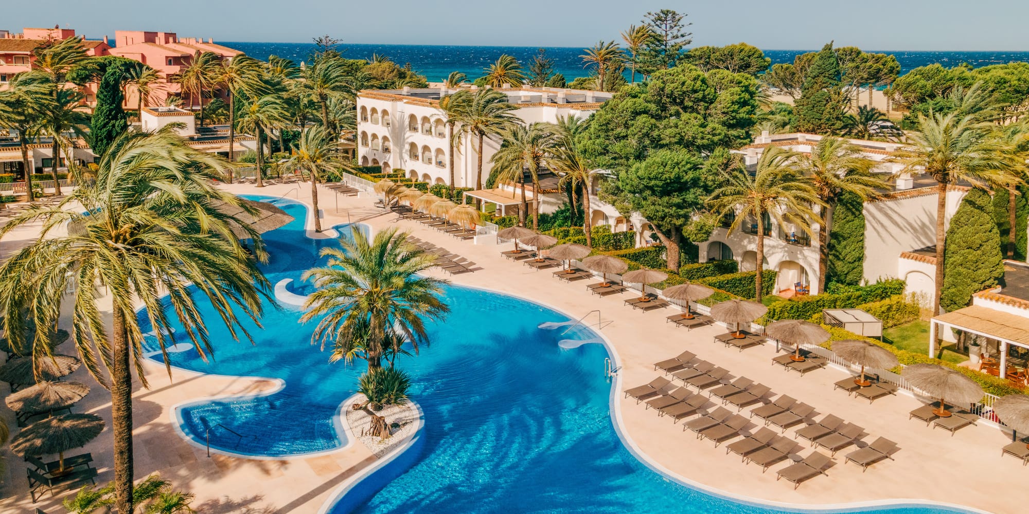 Tropical resort aerial with large blue pool, palm trees, sun loungers, and ocean.