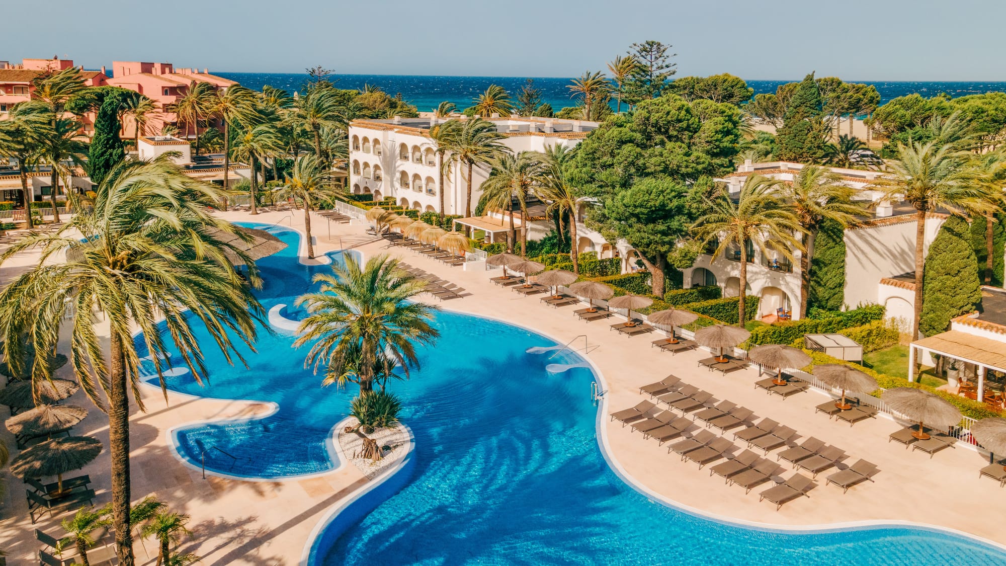 Resort hotel with winding blue pool, palm trees, sun loungers, ocean view.