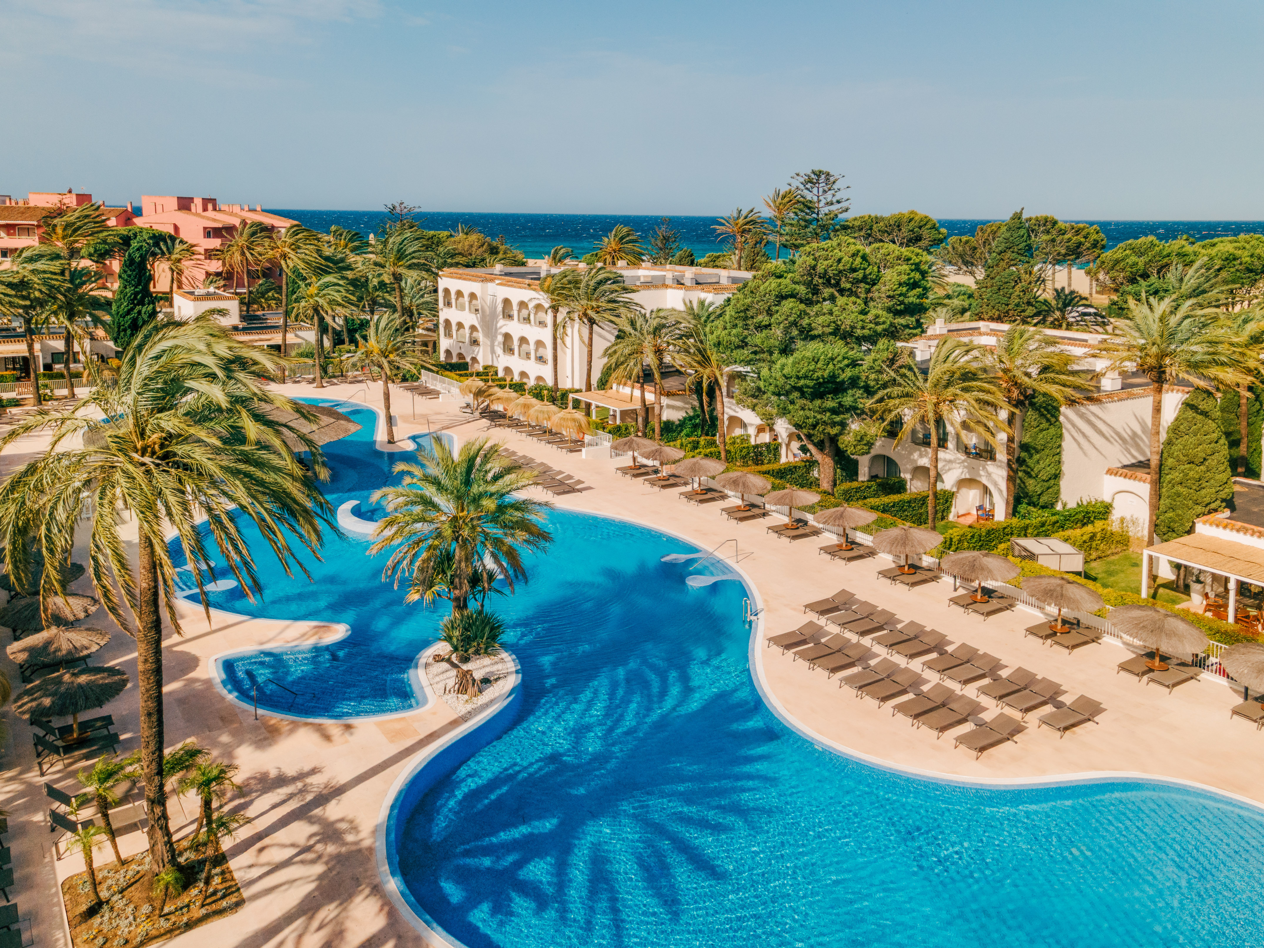 Tropical resort with large winding pool, palm trees, sunbeds, and ocean.