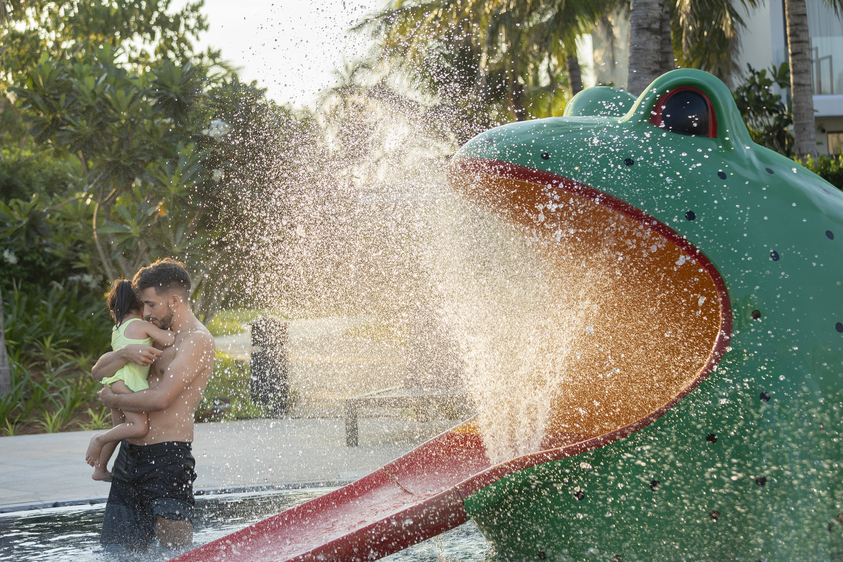 Father holds child near a large frog water slide splashing water in a pool.