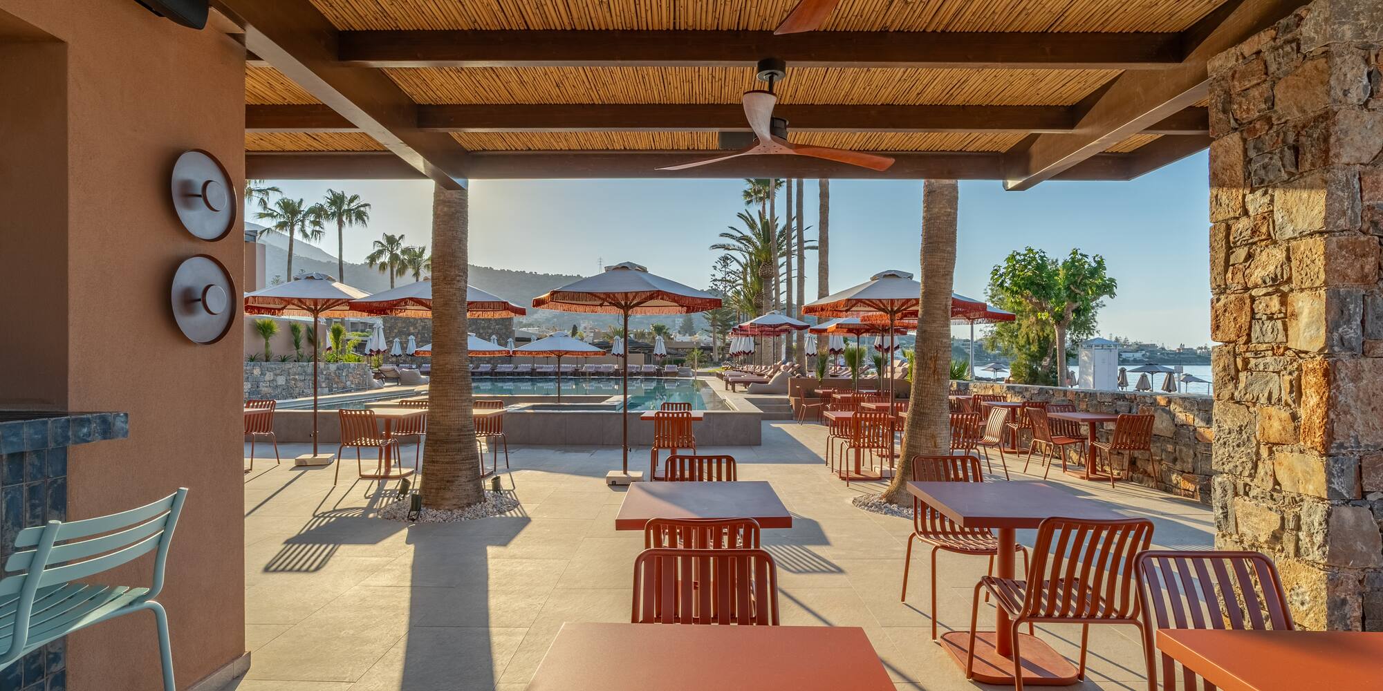 Resort poolside dining patio with terracotta tables, palm trees, and sea.