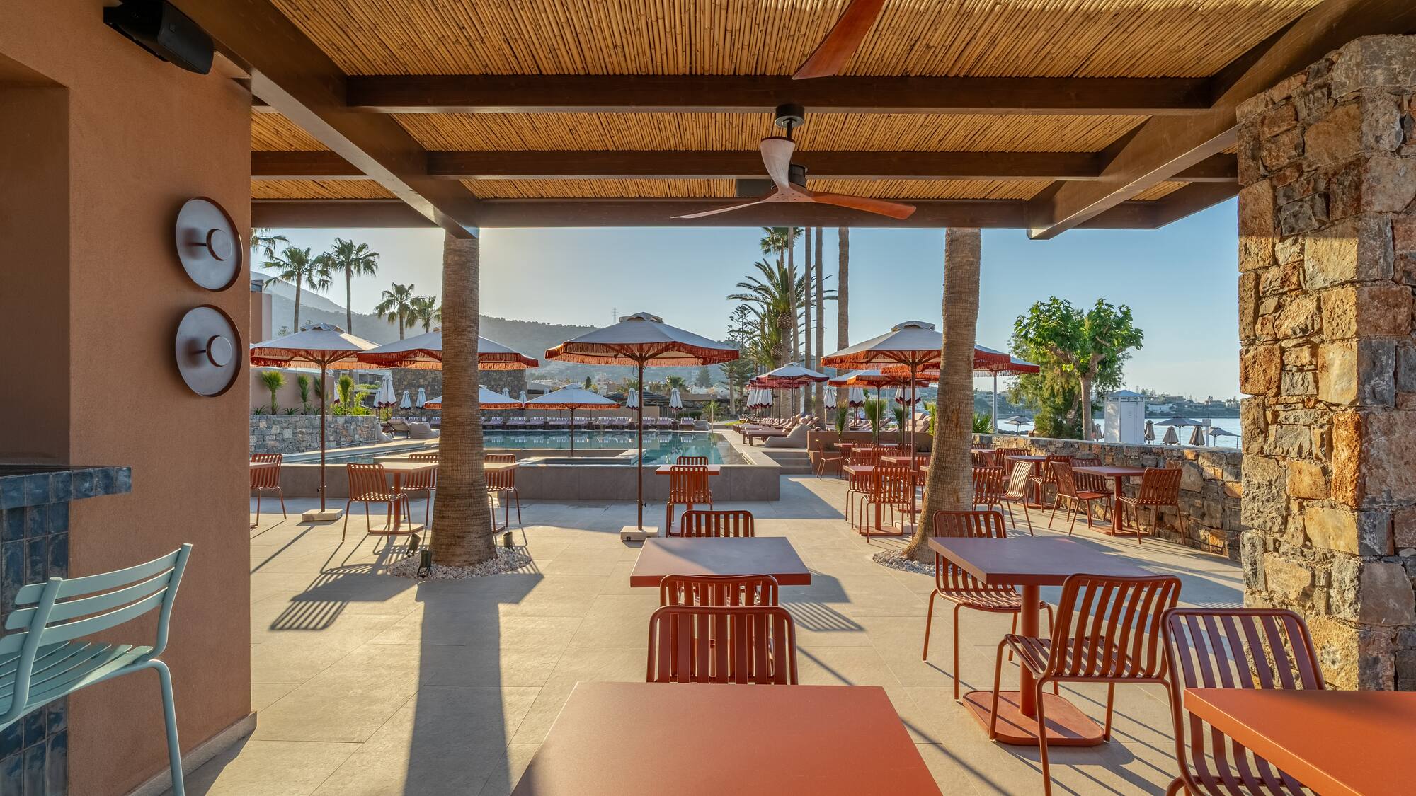 Resort poolside dining patio with terracotta tables, palm trees, and sea.