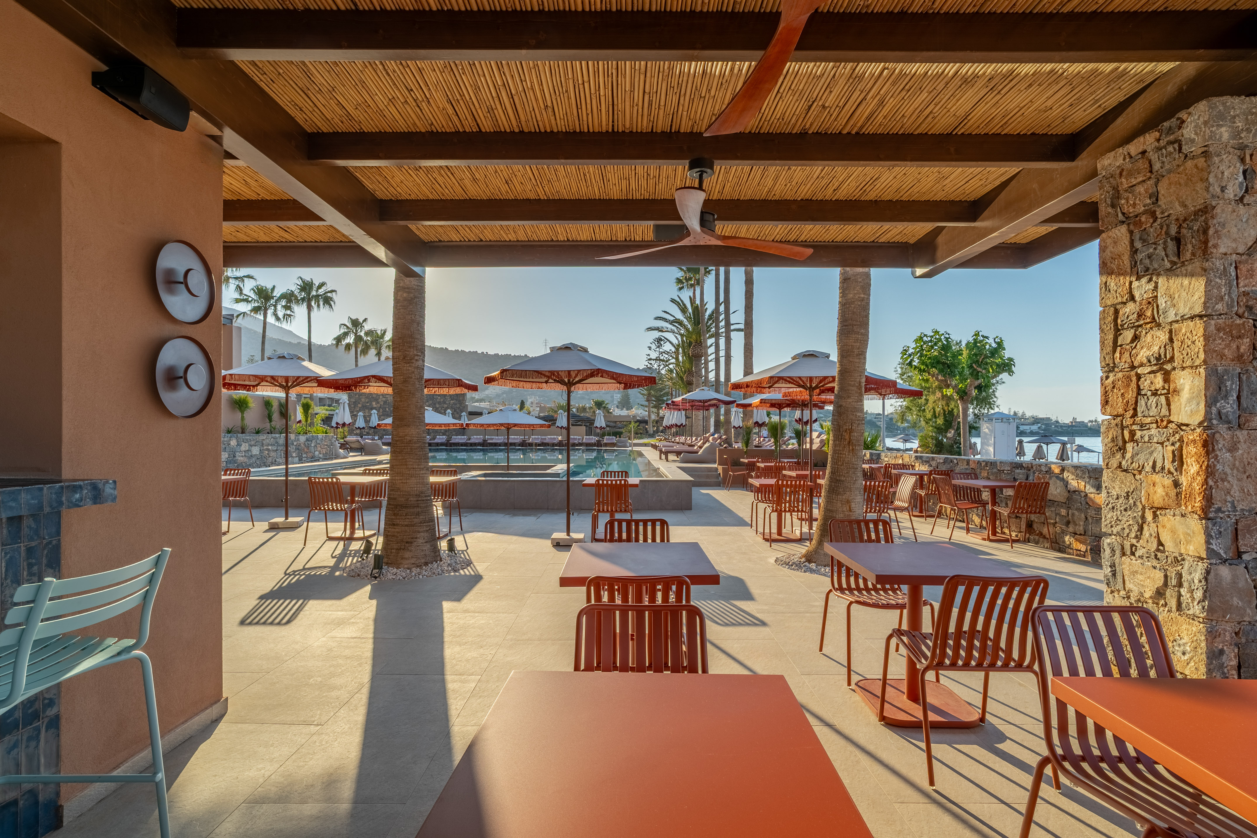 Resort poolside dining patio with terracotta tables, palm trees, and sea.