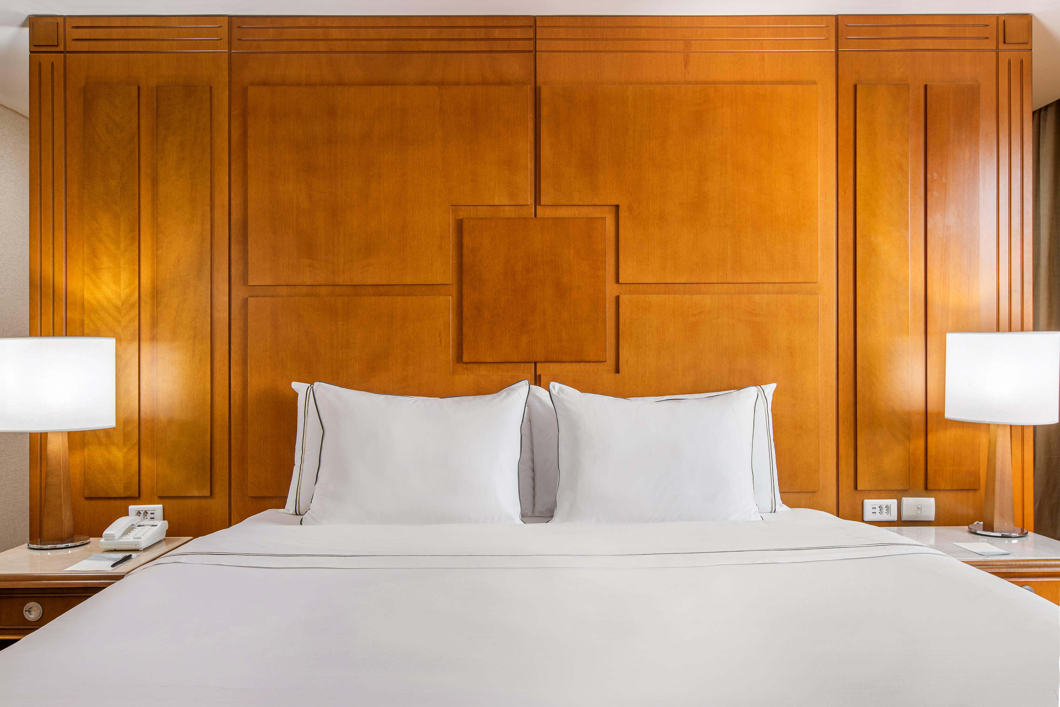 Hotel room with crisp white bed, pillows, wooden headboard, and bedside lamps.