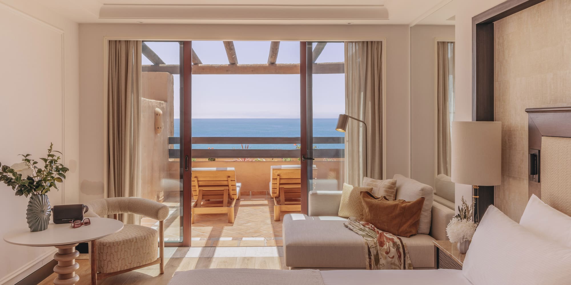 Ocean view hotel room with balcony, outdoor lounge chairs, bed, and sofa.