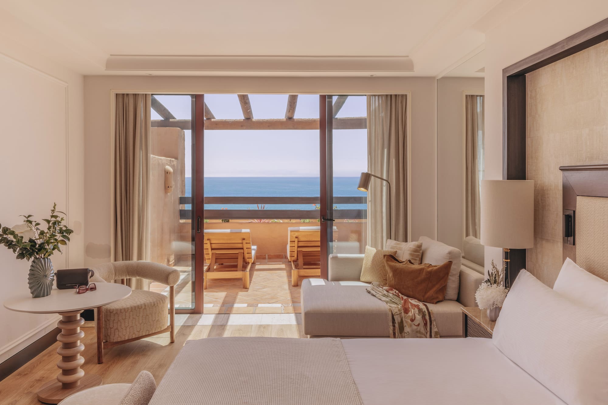 Ocean view hotel room with balcony, outdoor lounge chairs, bed, and sofa.