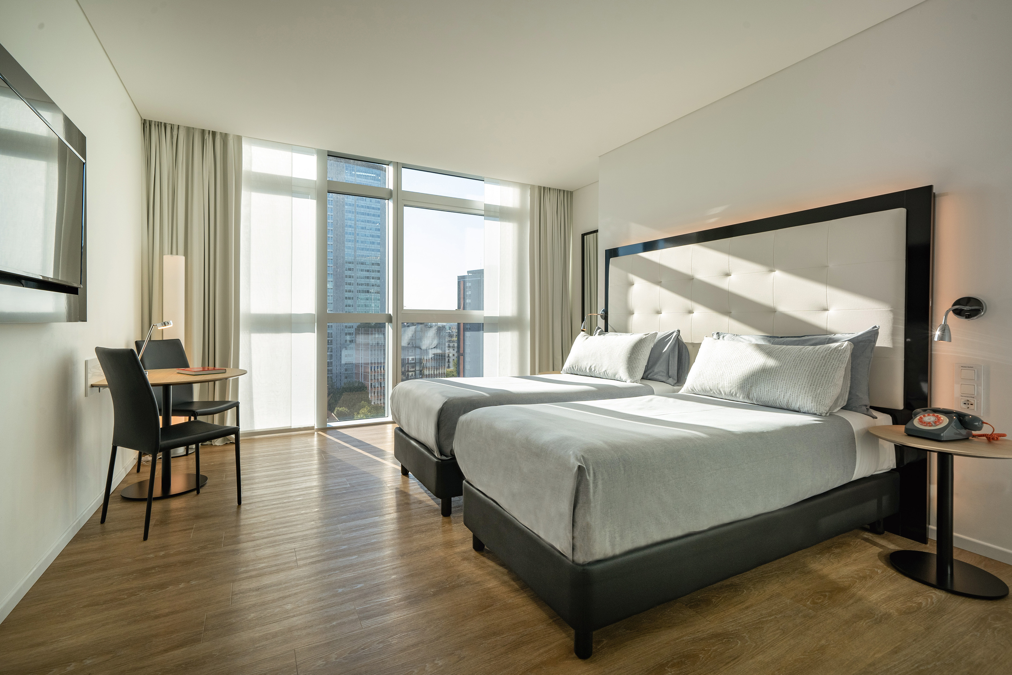 Hotel room with twin beds, city view, light wood floor, and workspace.