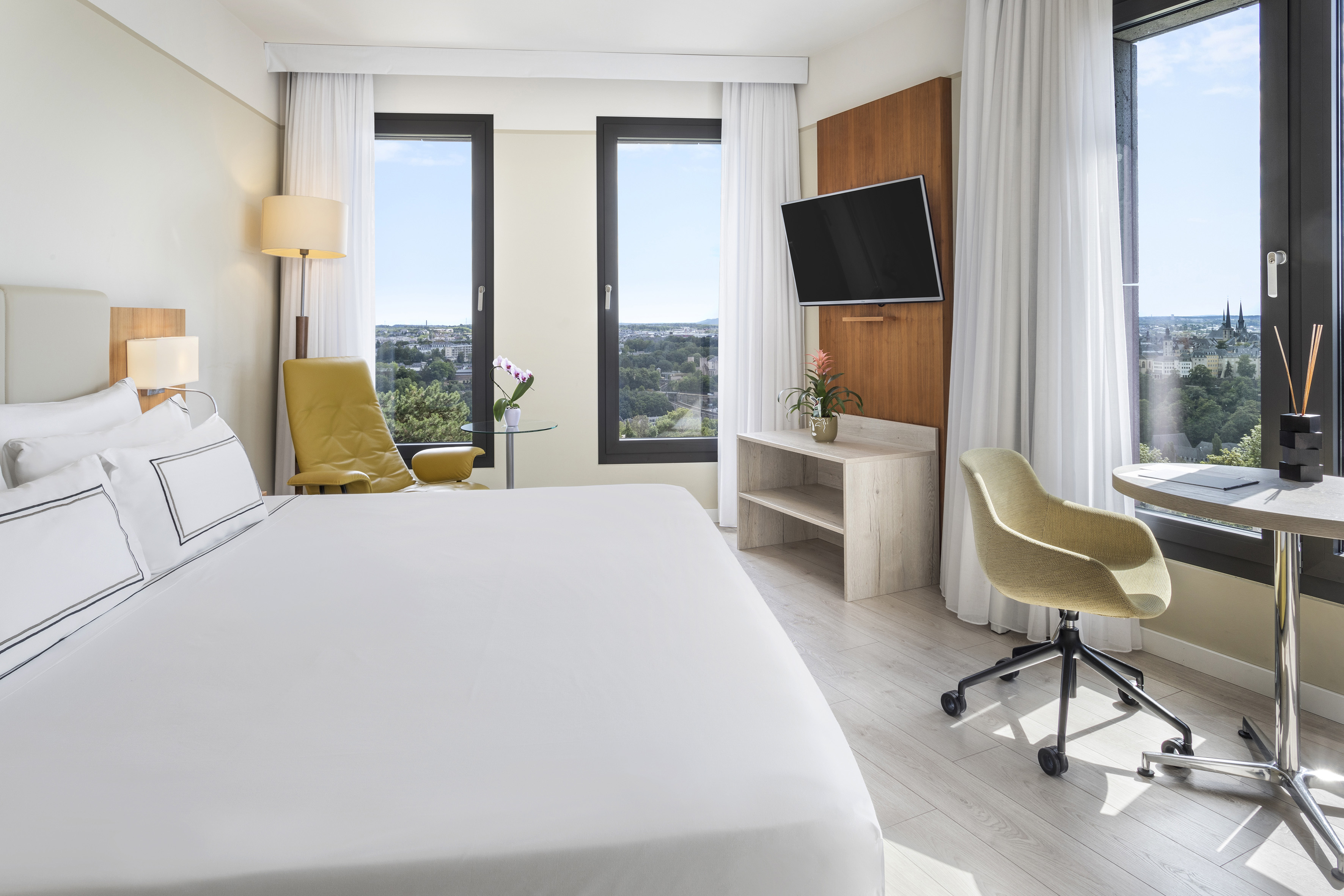 Modern hotel room with large bed, scenic city view windows, TV, and work desk.