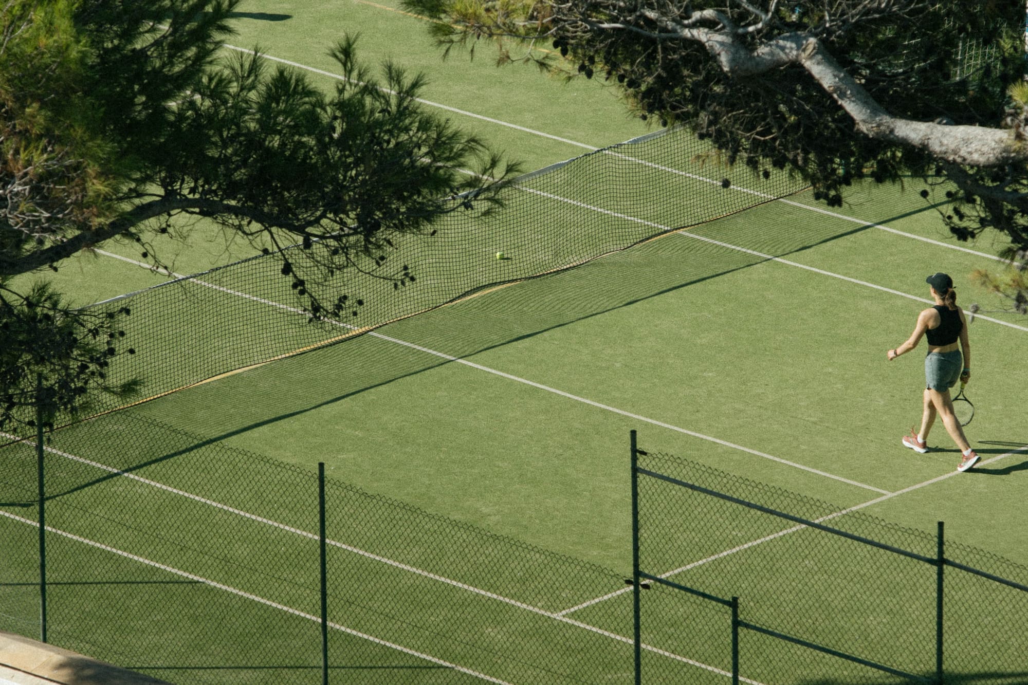 Mujer caminando en cancha de tenis verde con raqueta bajo pinos.