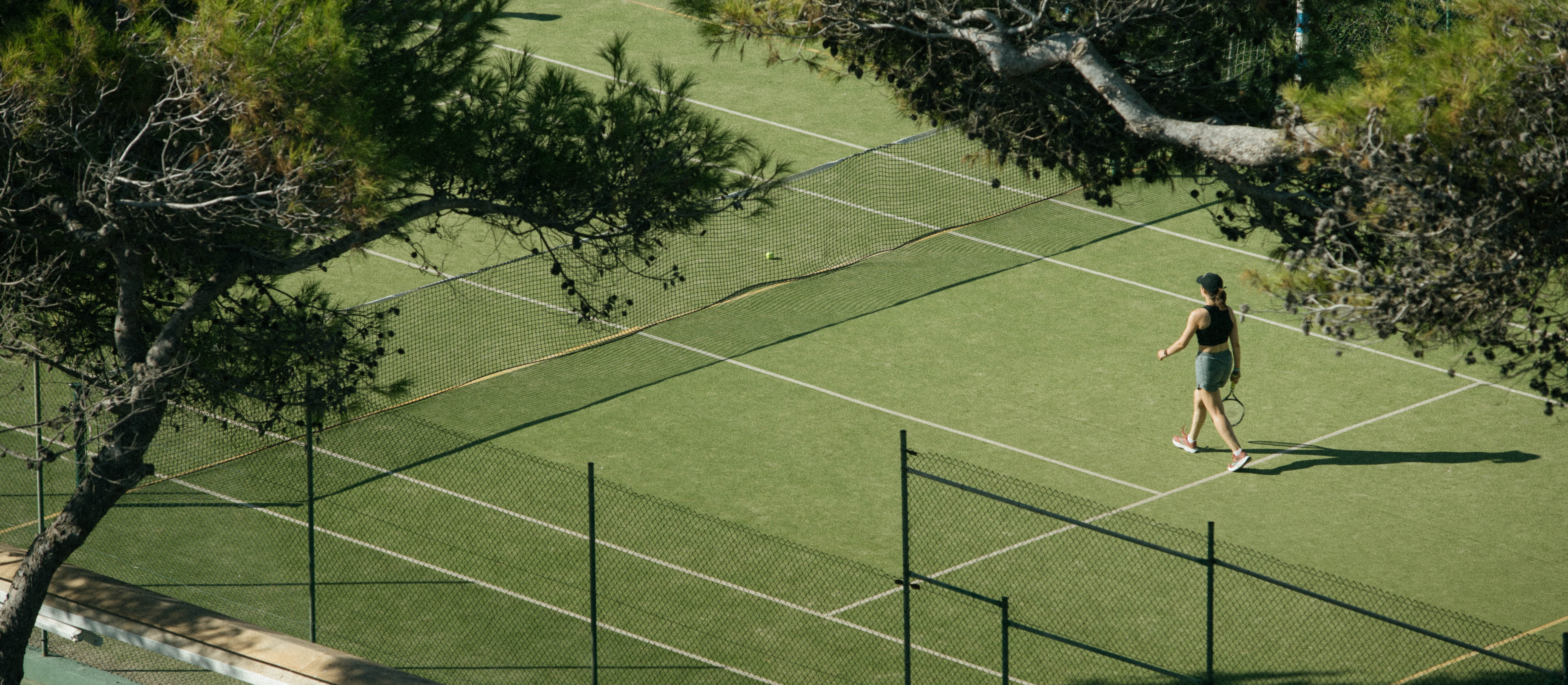 Mujer caminando en cancha de tenis verde con raqueta bajo pinos.