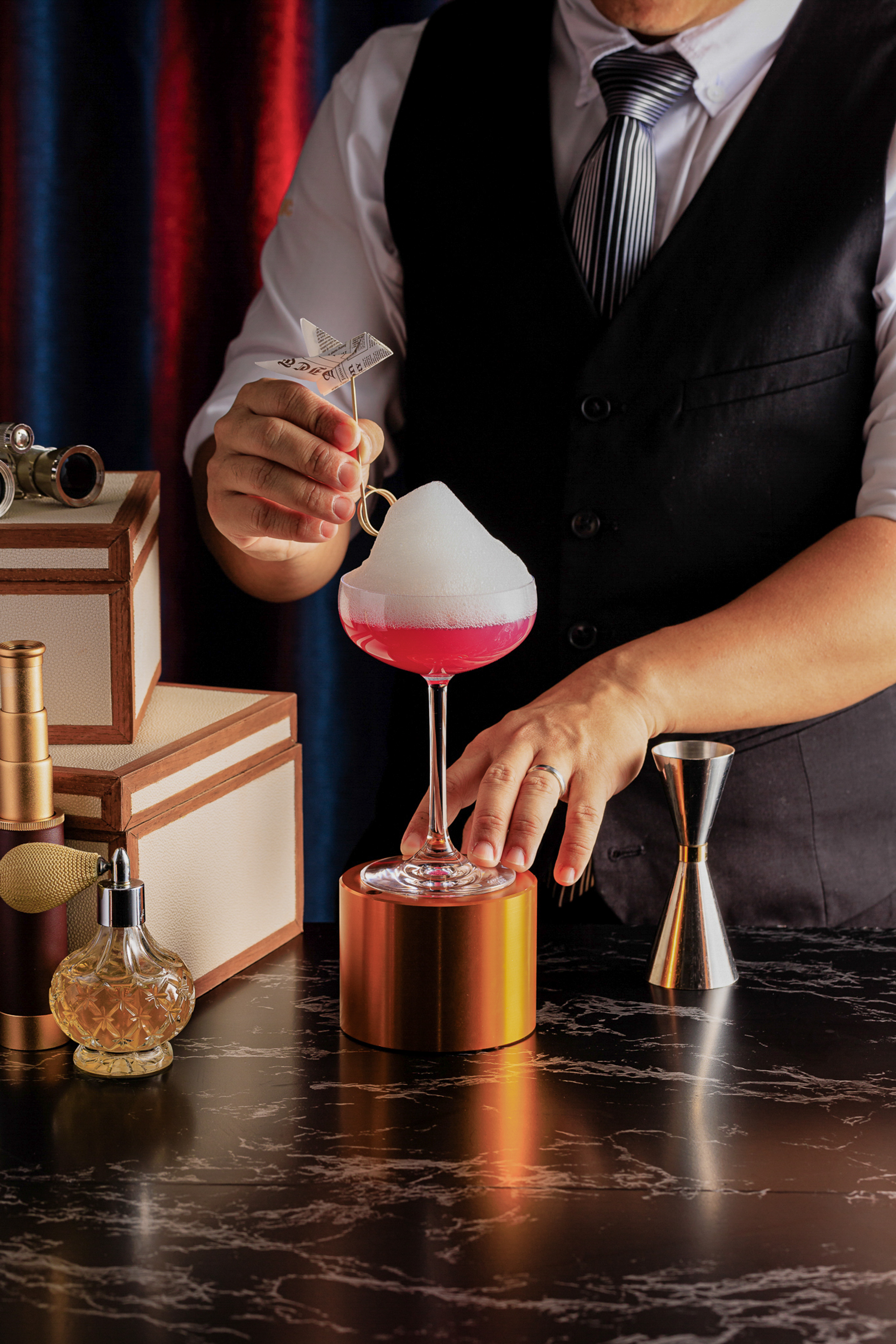 Bartender garnishing a red foamy cocktail with a paper airplane at a stylish bar.