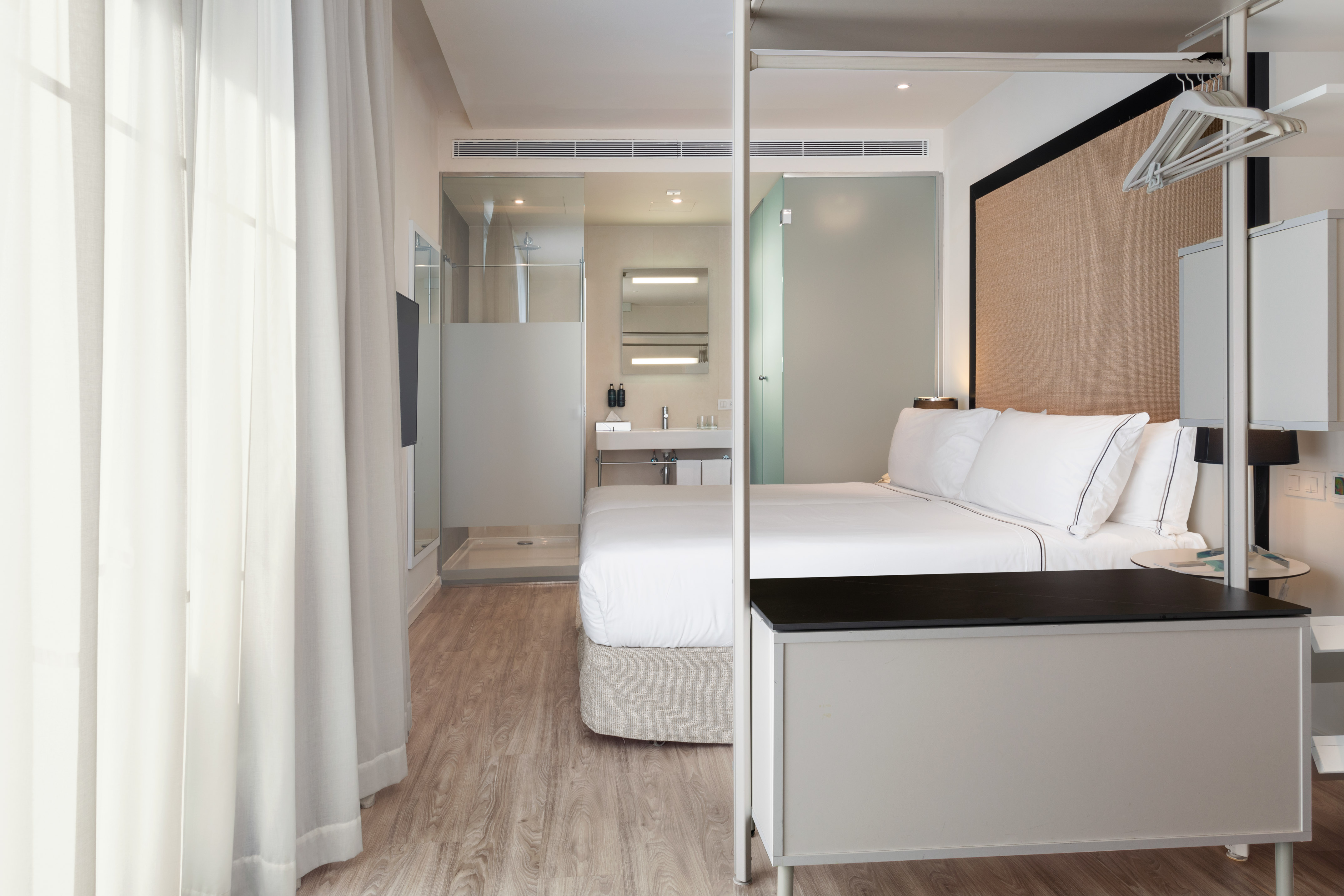 Modern hotel bedroom with white bed, open shower bathroom, and wood flooring.