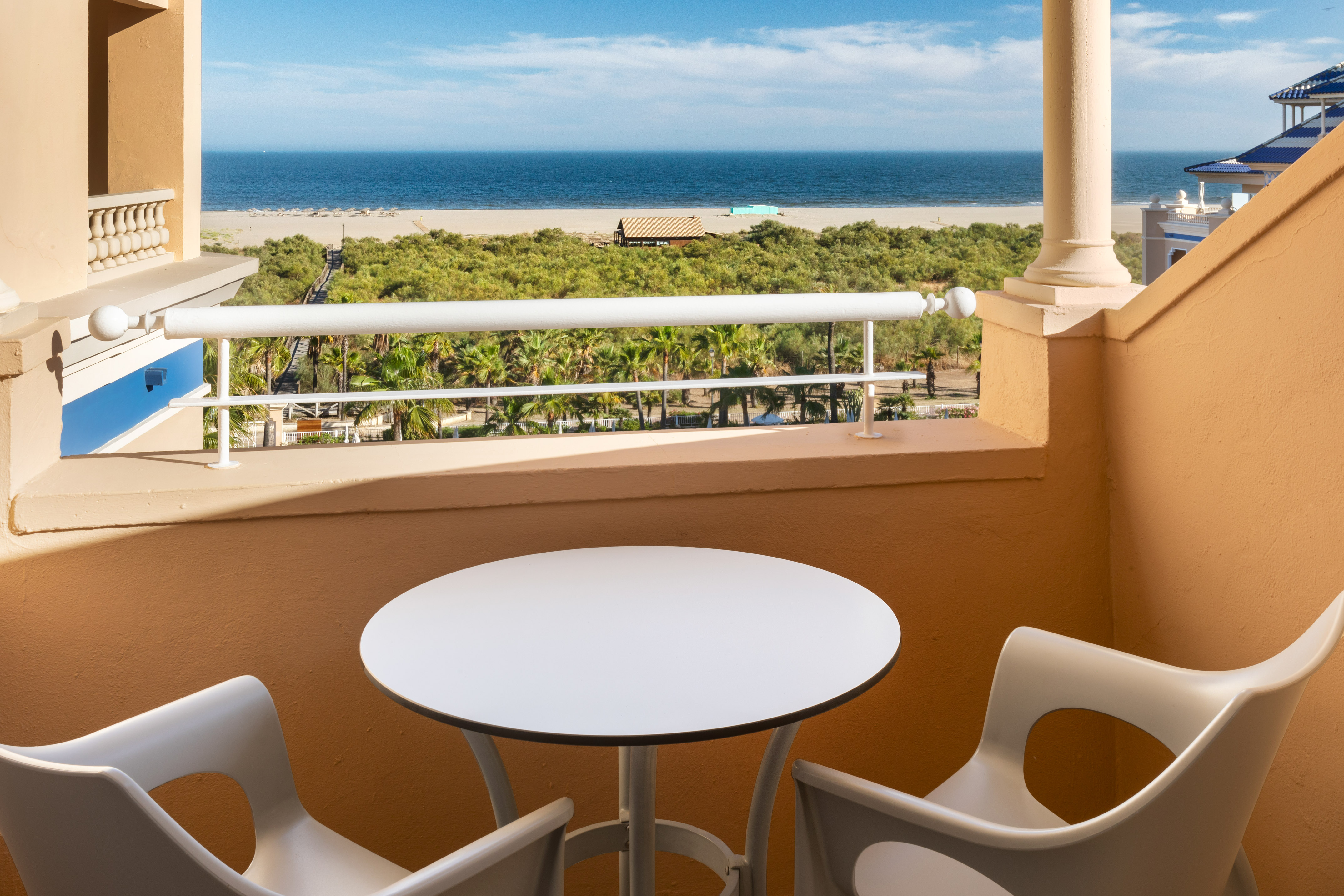 Resort balcony overlooking sandy beach, blue ocean, and tropical palm trees.
