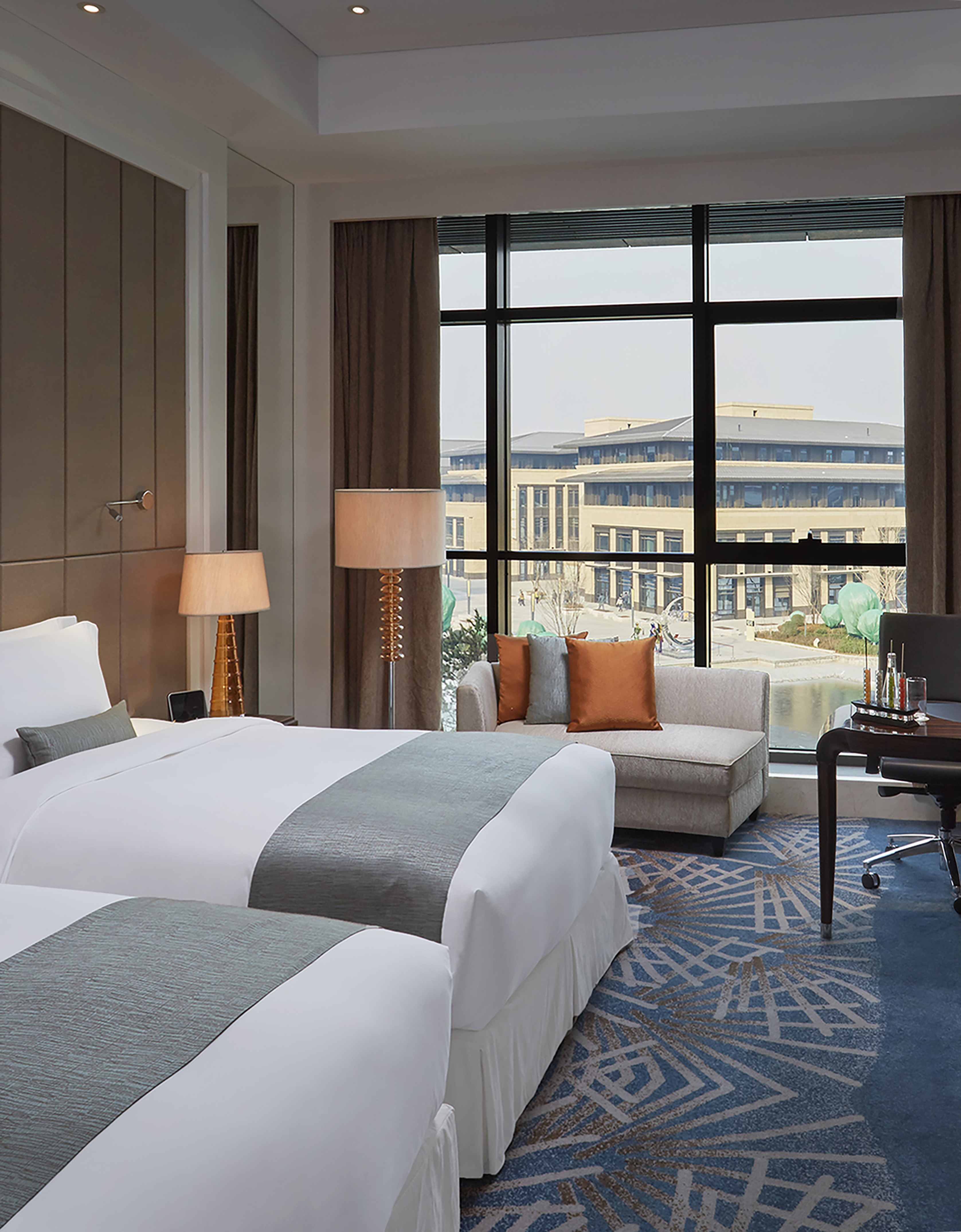 Modern hotel room with two beds, city view window, sofa, and desk.