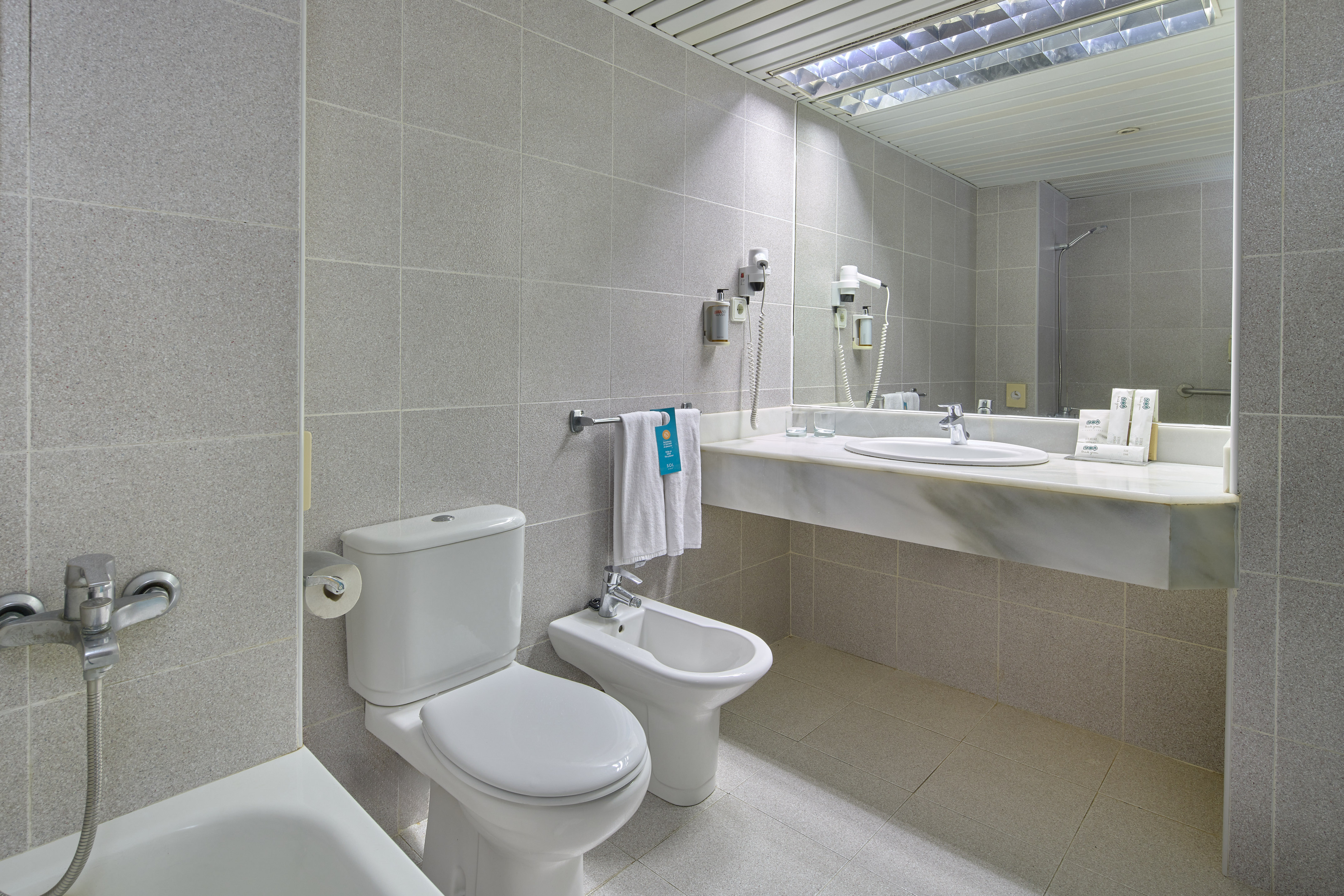 Modern grey tiled hotel bathroom with toilet, bidet, sink, mirror, and bathtub.
