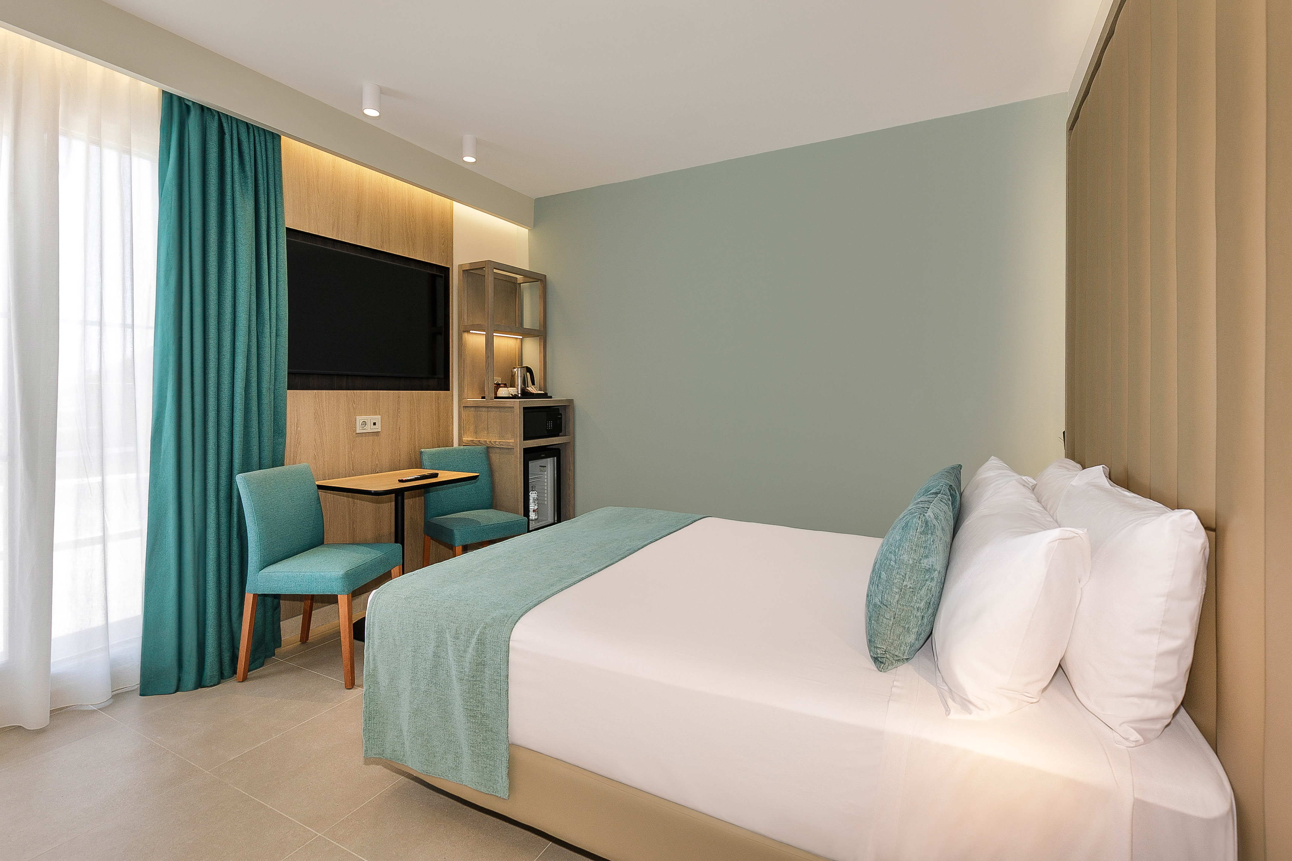 Modern hotel room with bed, teal decor, flat-screen TV, and small seating area.