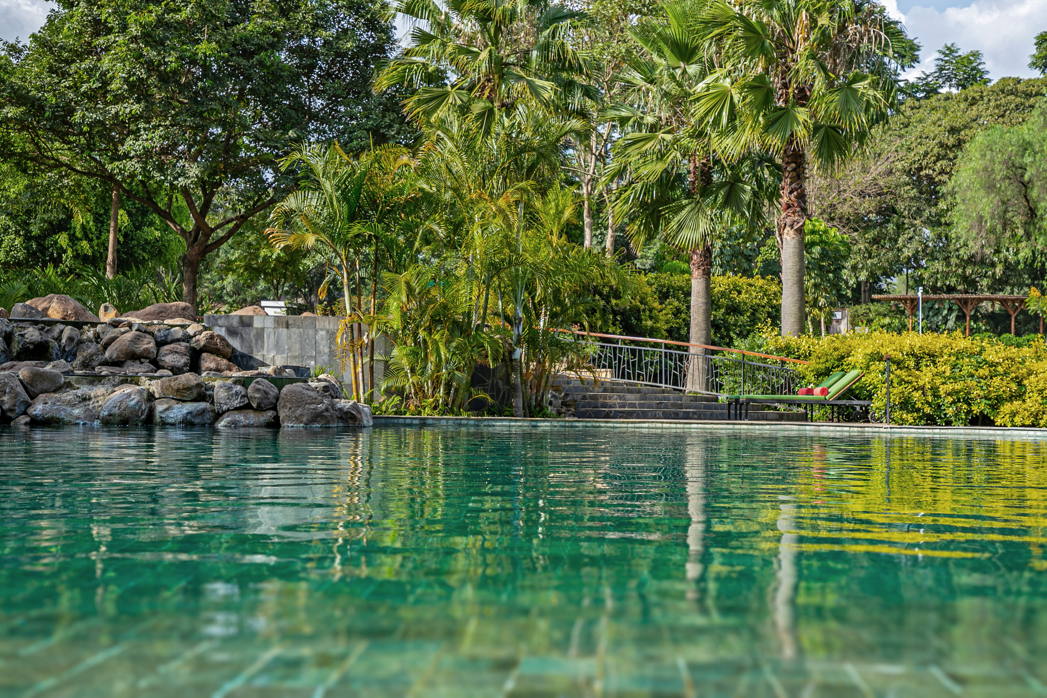Tropical resort pool with palm trees, rock feature, and lounge chair.
