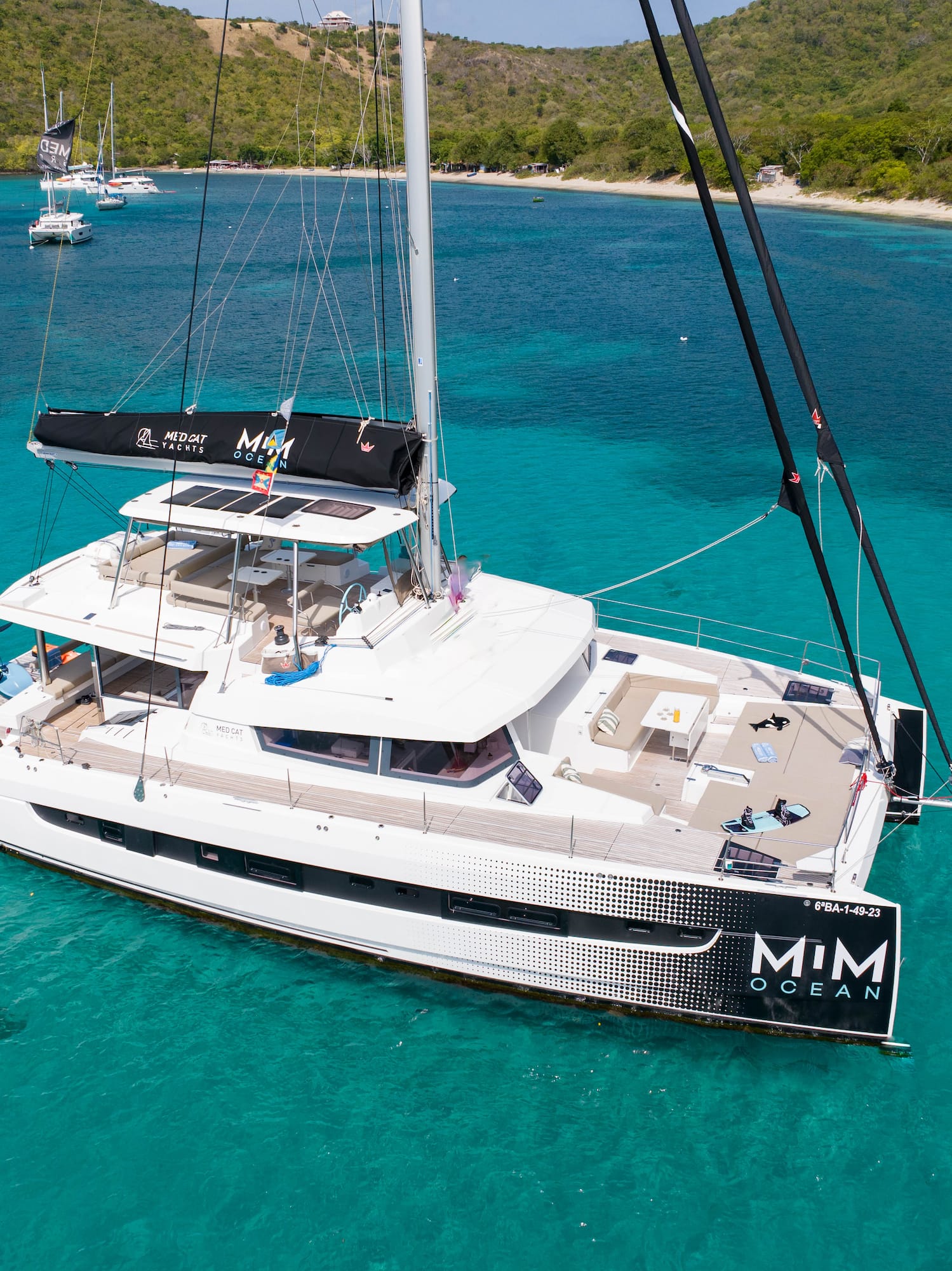 White MIM OCEAN catamaran yacht anchored in clear turquoise bay, tropical hills.