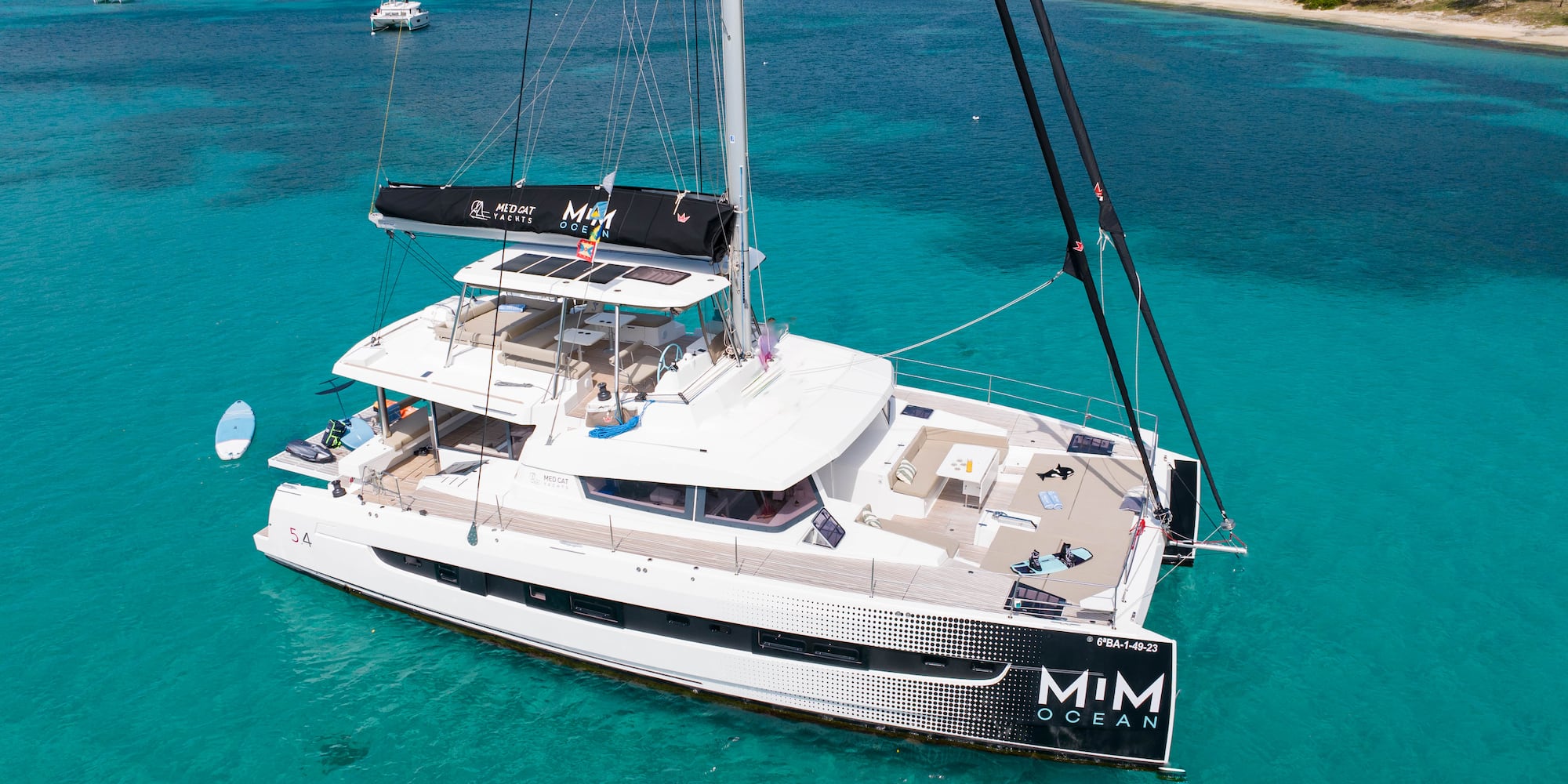 White MIM OCEAN catamaran yacht anchored in clear turquoise bay, tropical hills.