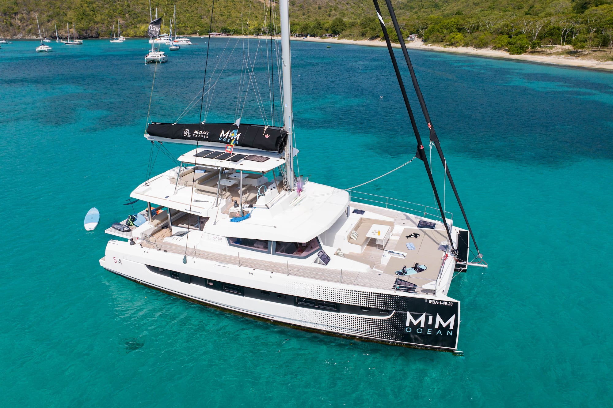 White MIM OCEAN catamaran yacht anchored in clear turquoise bay, tropical hills.