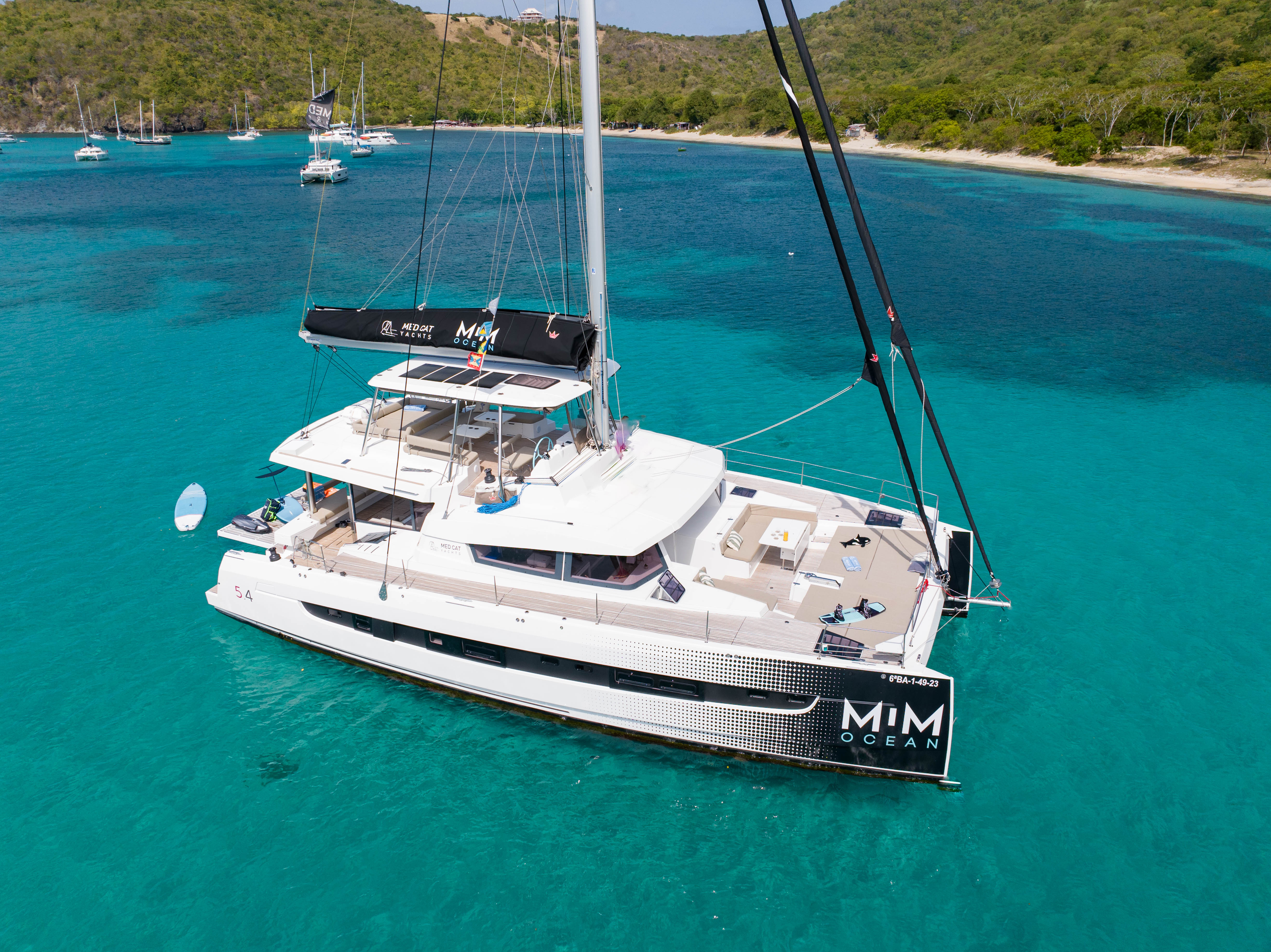 White MIM OCEAN catamaran yacht anchored in clear turquoise bay, tropical hills.