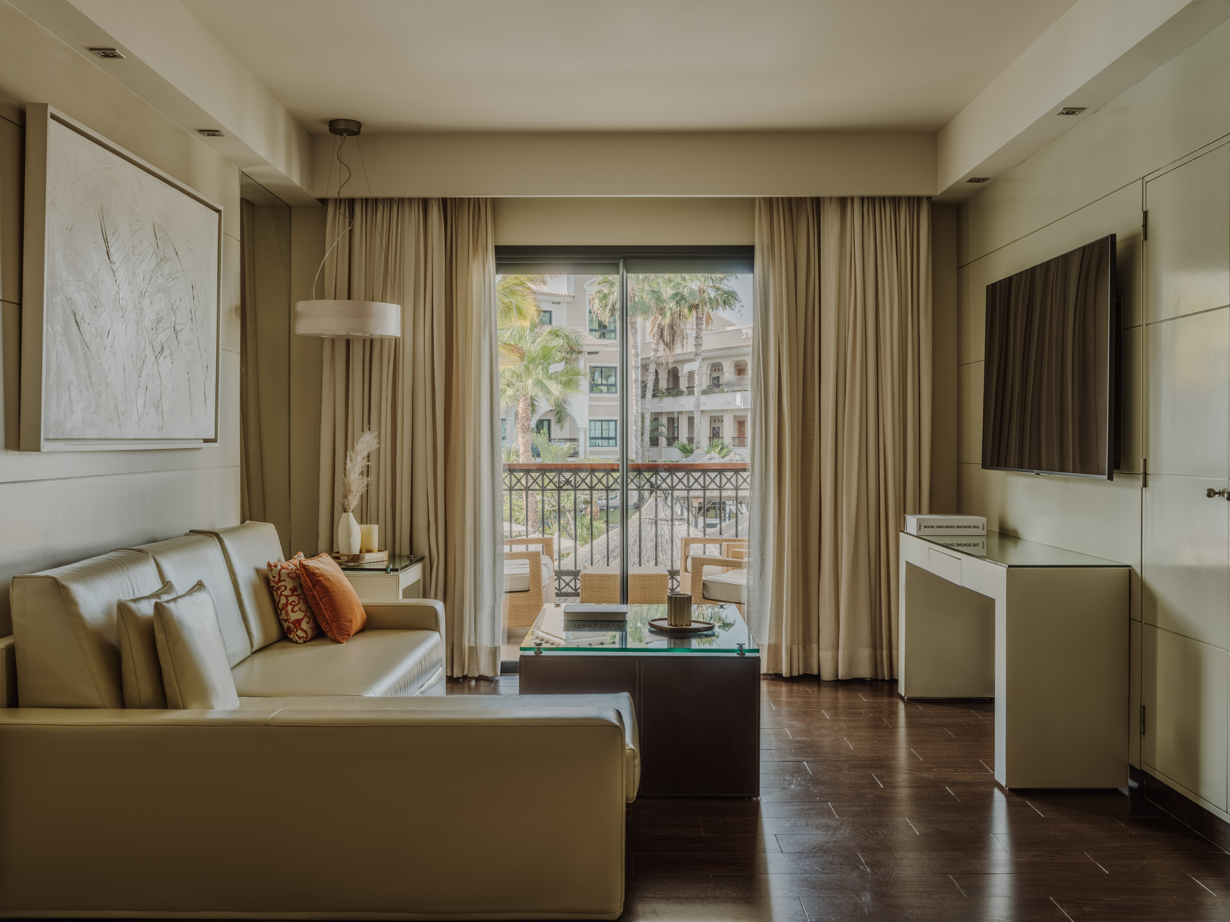 Hotel suite living room, cream sofa, balcony with resort palm tree view.