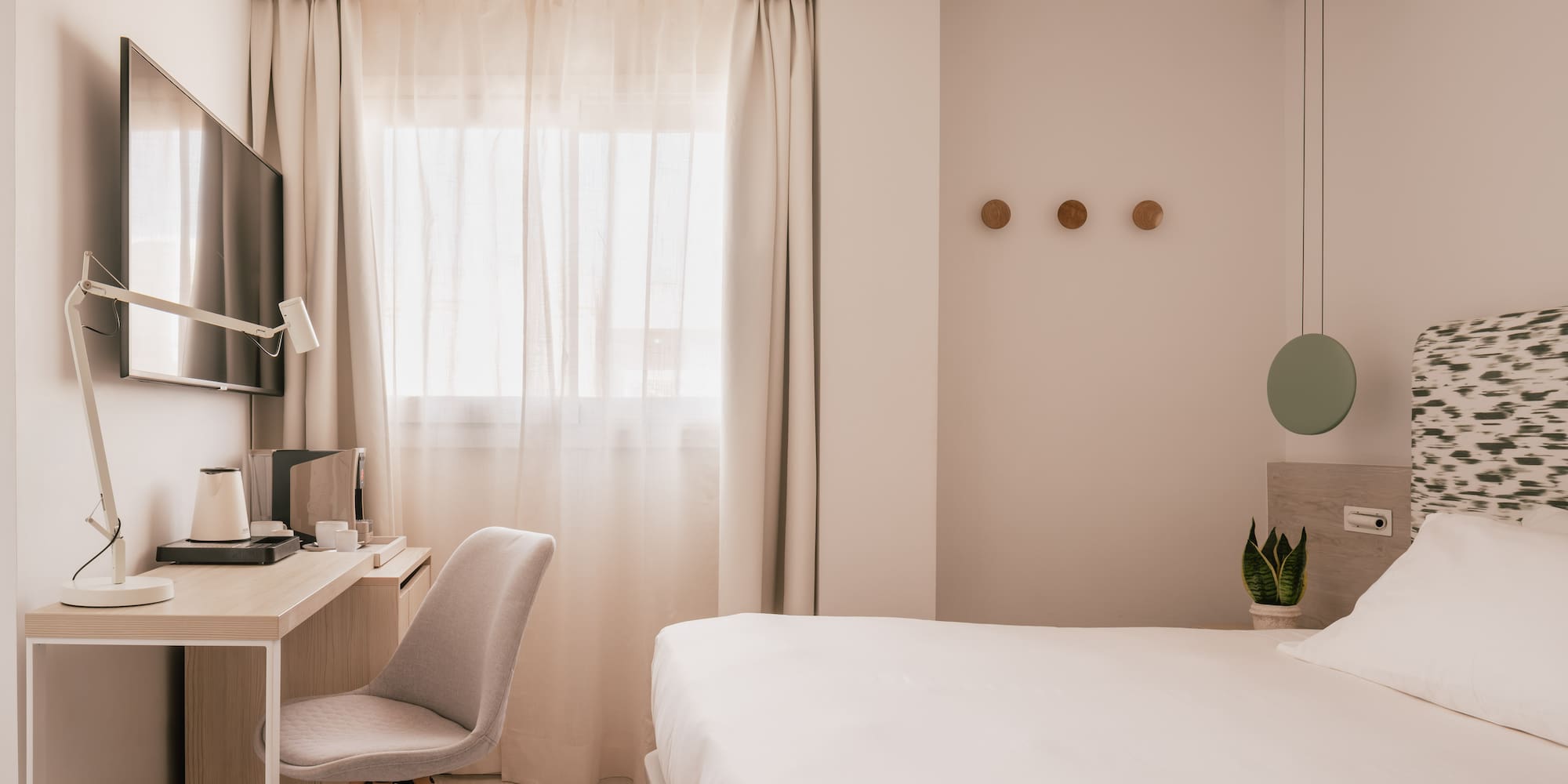 Modern hotel room with white bed, desk, TV, and window with sheer curtains.