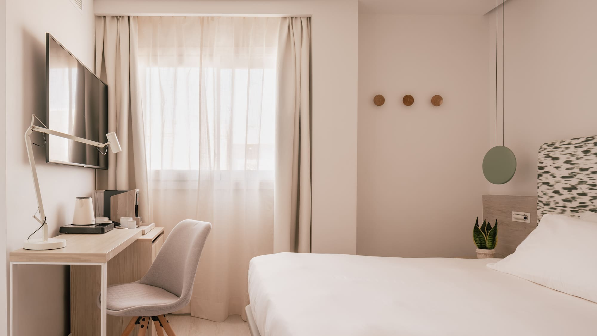 Modern hotel room with white bed, desk, TV, and window with sheer curtains.