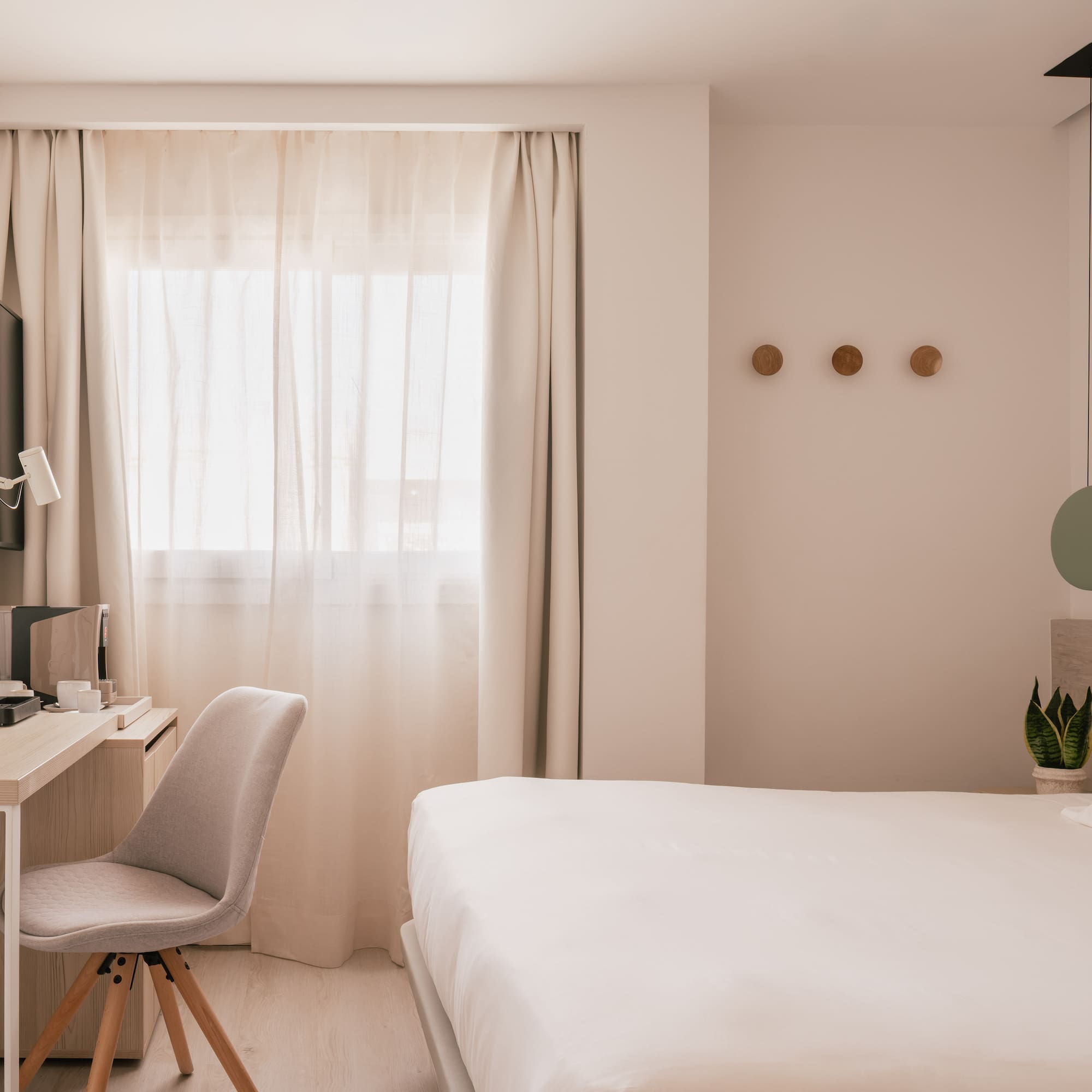 Modern hotel room with white bed, desk, TV, and window with sheer curtains.