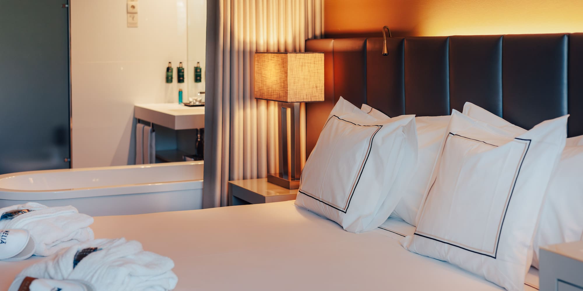 Elegant Meliá hotel room with bed, warm lamp, robes, and visible bathtub.
