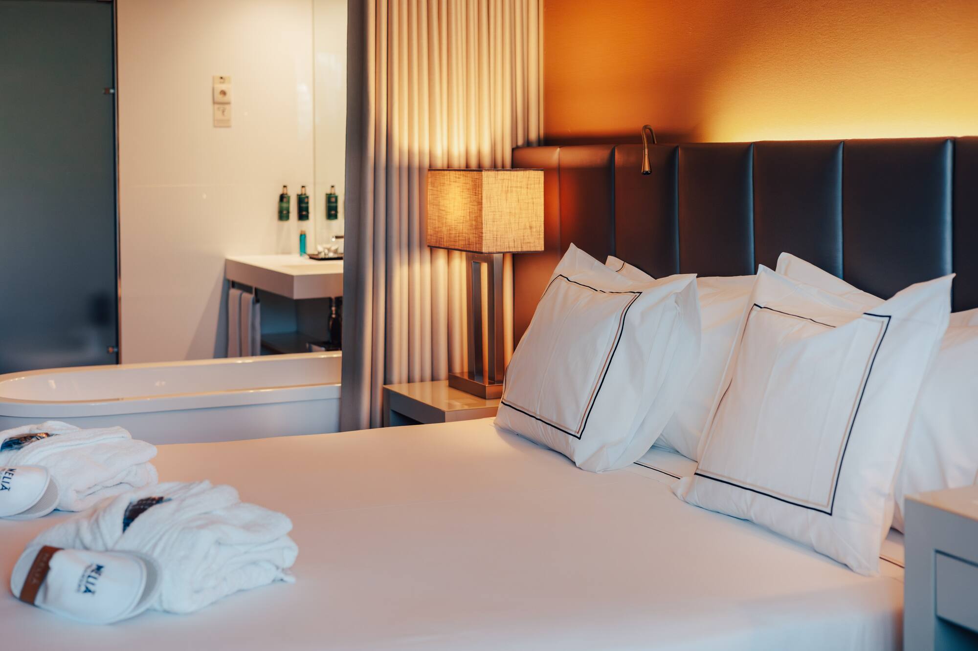 Elegant Meliá hotel room with bed, warm lamp, robes, and visible bathtub.