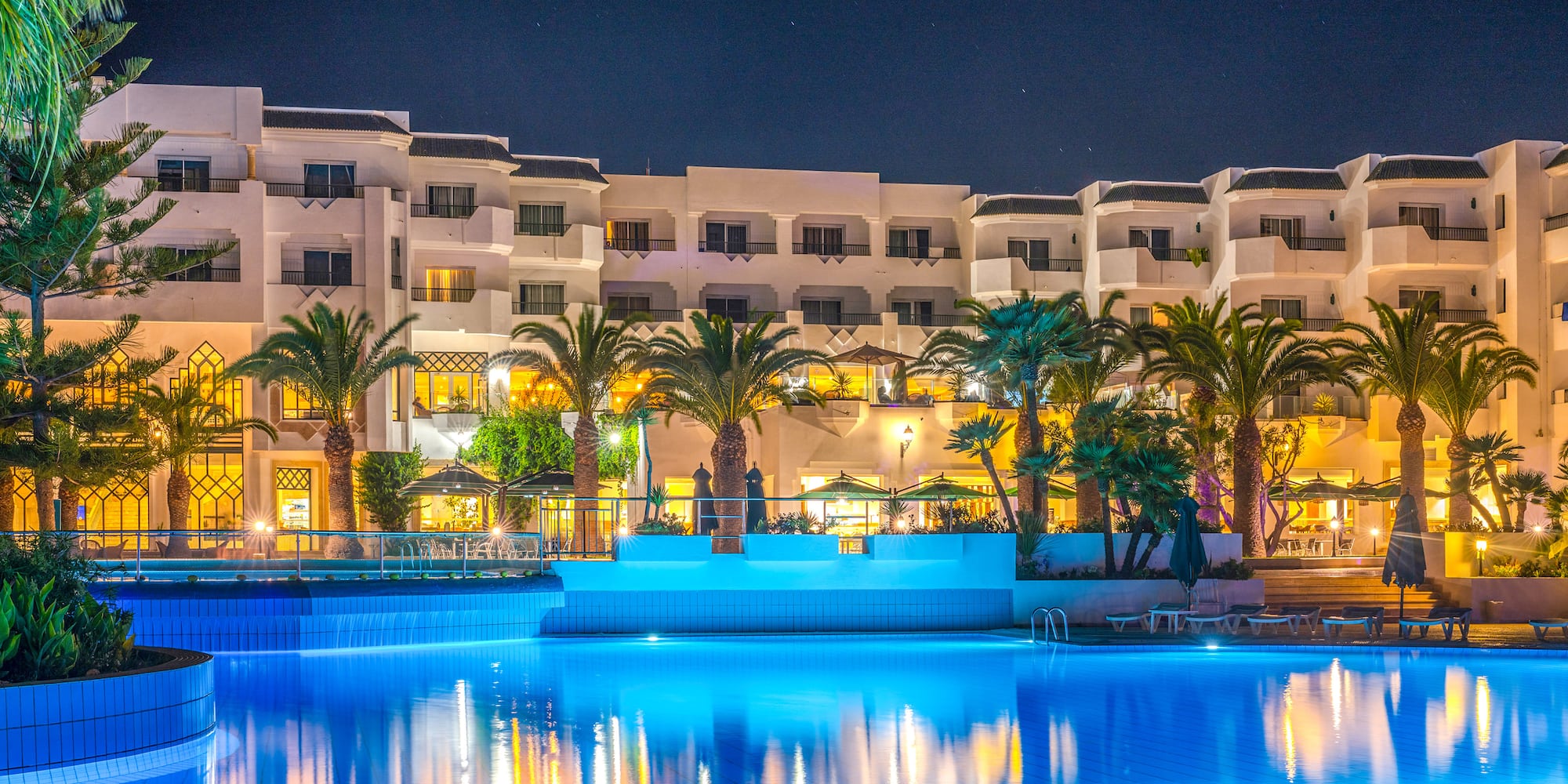 Illuminated hotel resort with a bright blue swimming pool and palm trees at night.