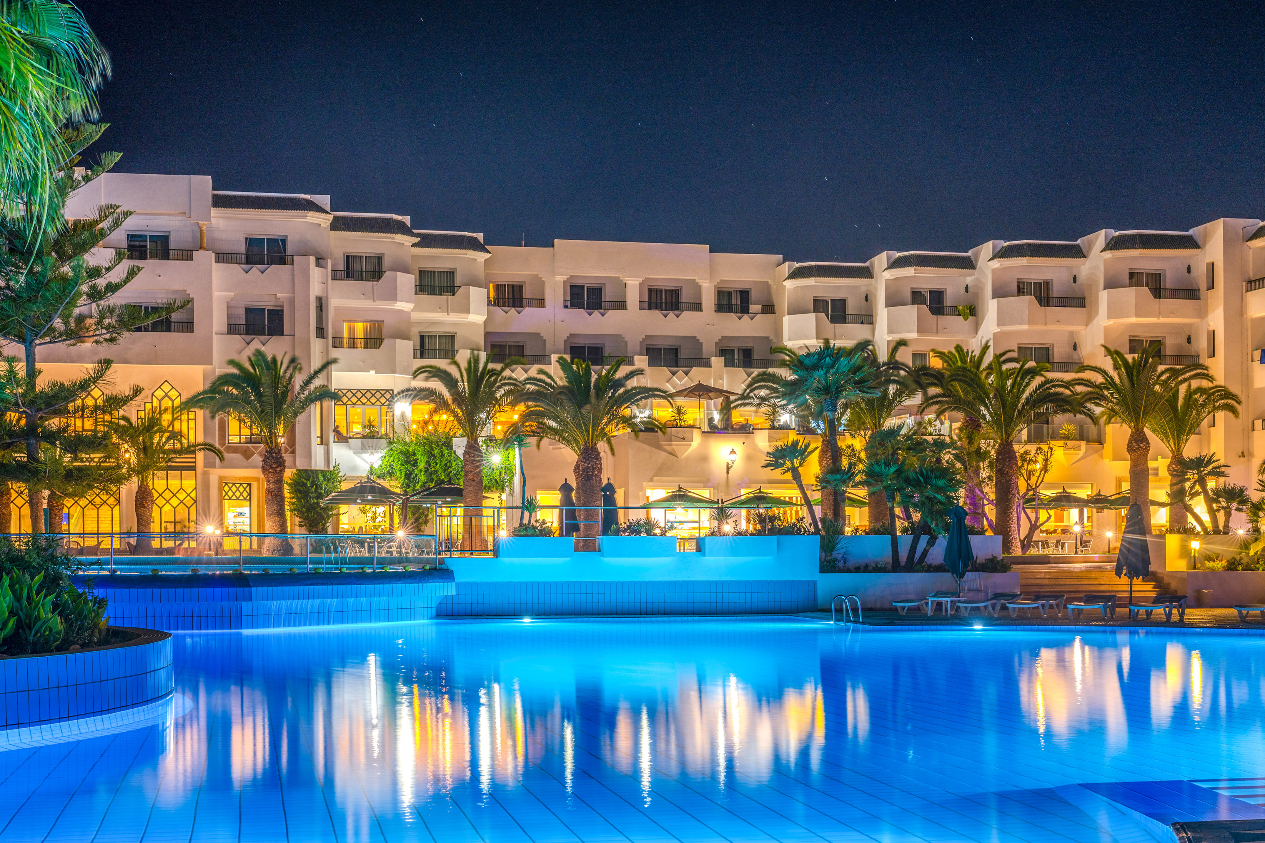 Illuminated hotel resort with a bright blue swimming pool and palm trees at night.