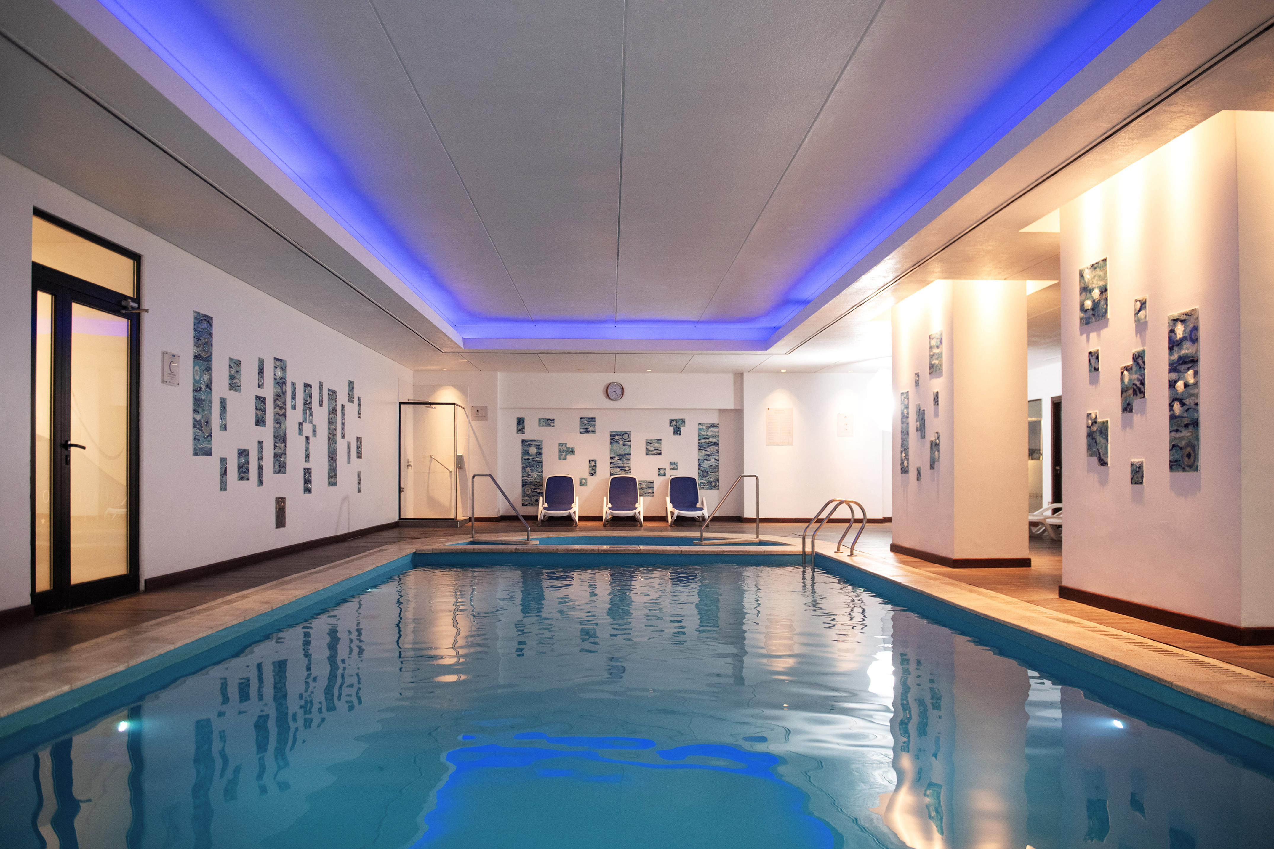 Indoor pool with blue ceiling light, abstract walls, and three lounge chairs.