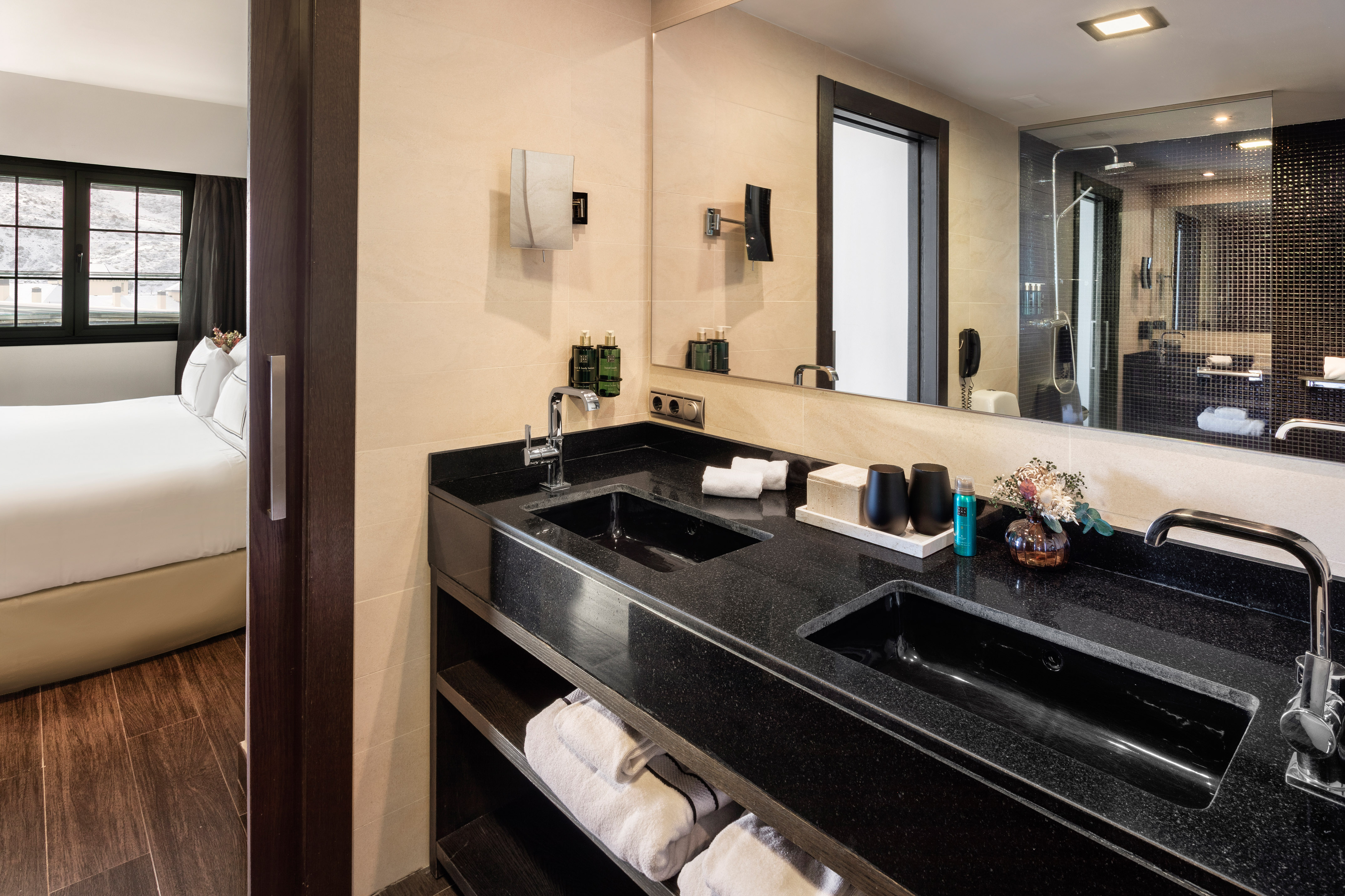 Hotel bathroom with black granite double vanity, mirror, and connected bedroom.