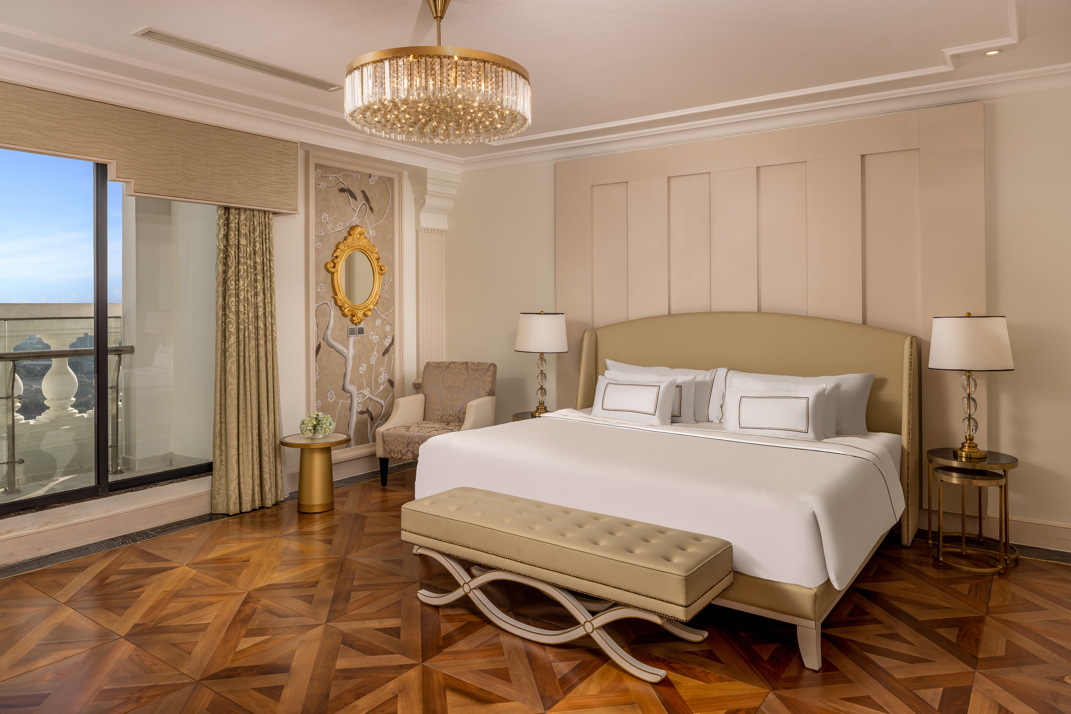 Elegant hotel bedroom with king bed, chandelier, wood floor, and balcony view.