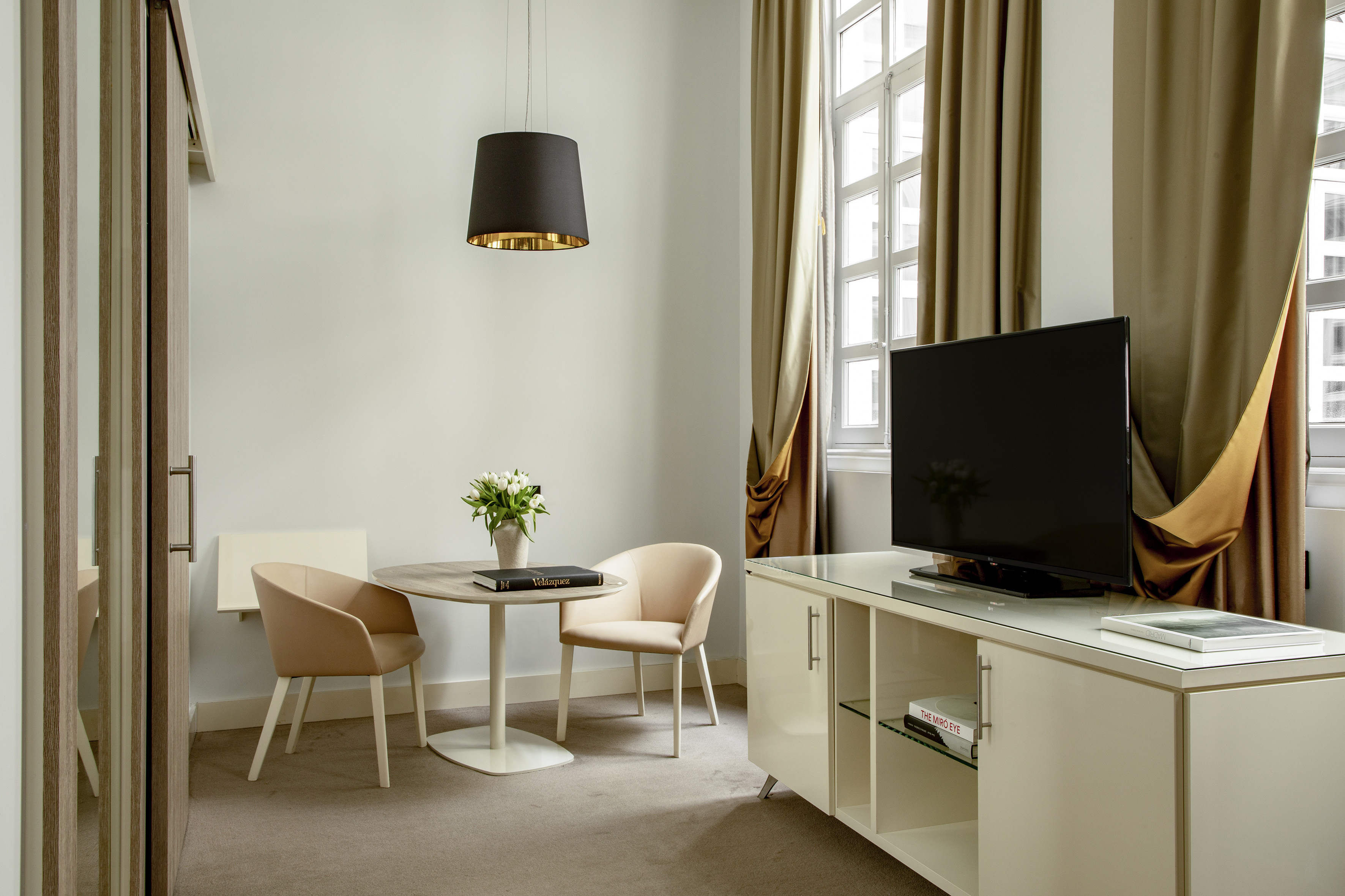 Modern hotel suite living area with dining table, chairs, TV, and window.