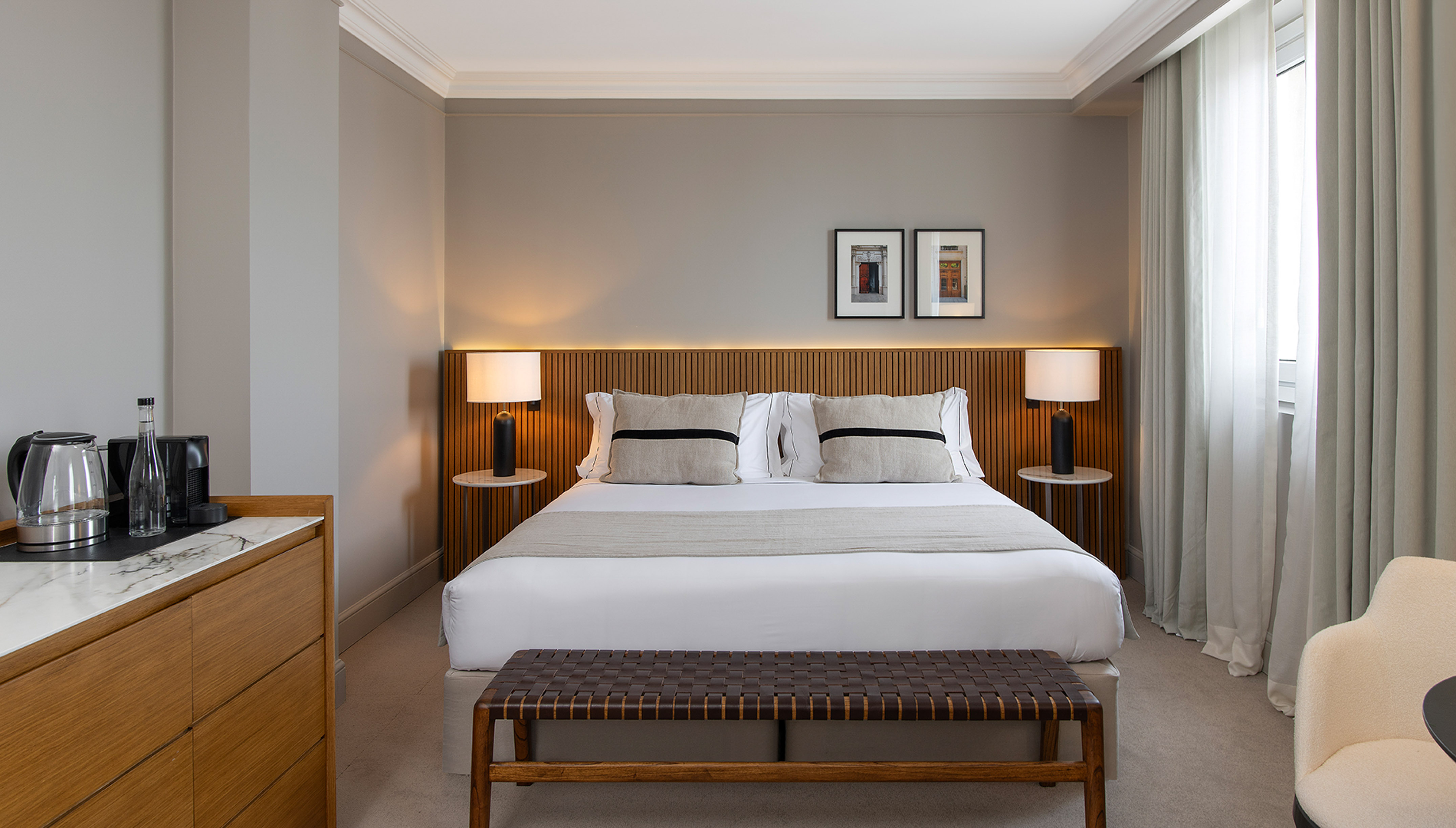 Modern hotel bedroom with king bed, wood headboard, bedside lamps, and woven bench.